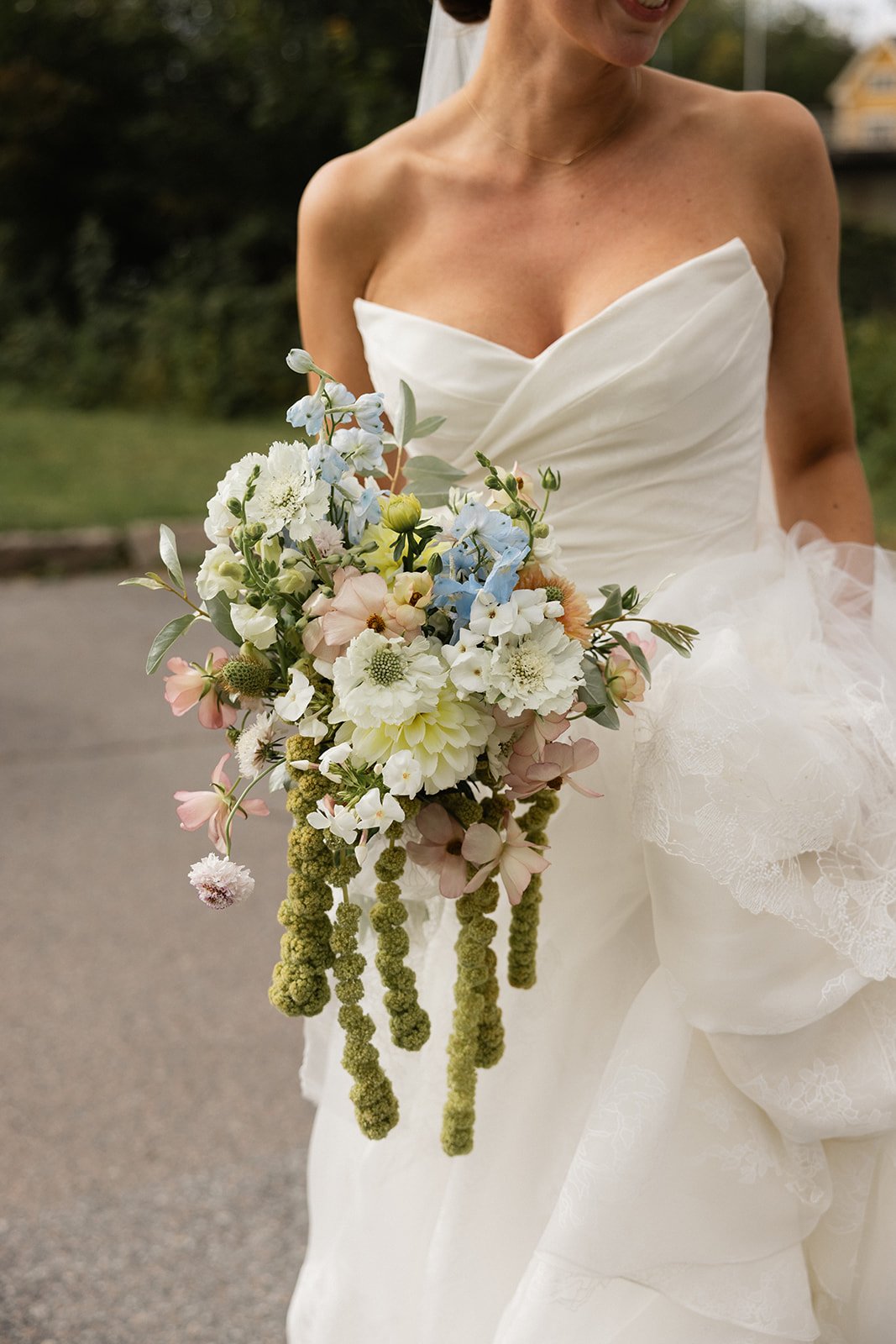 Nærbilde av en brud som holder en blomsterbuket, som består av ulike hvite, rosa, blå blomster og grønne blader, på en utendørs bakgrunn.