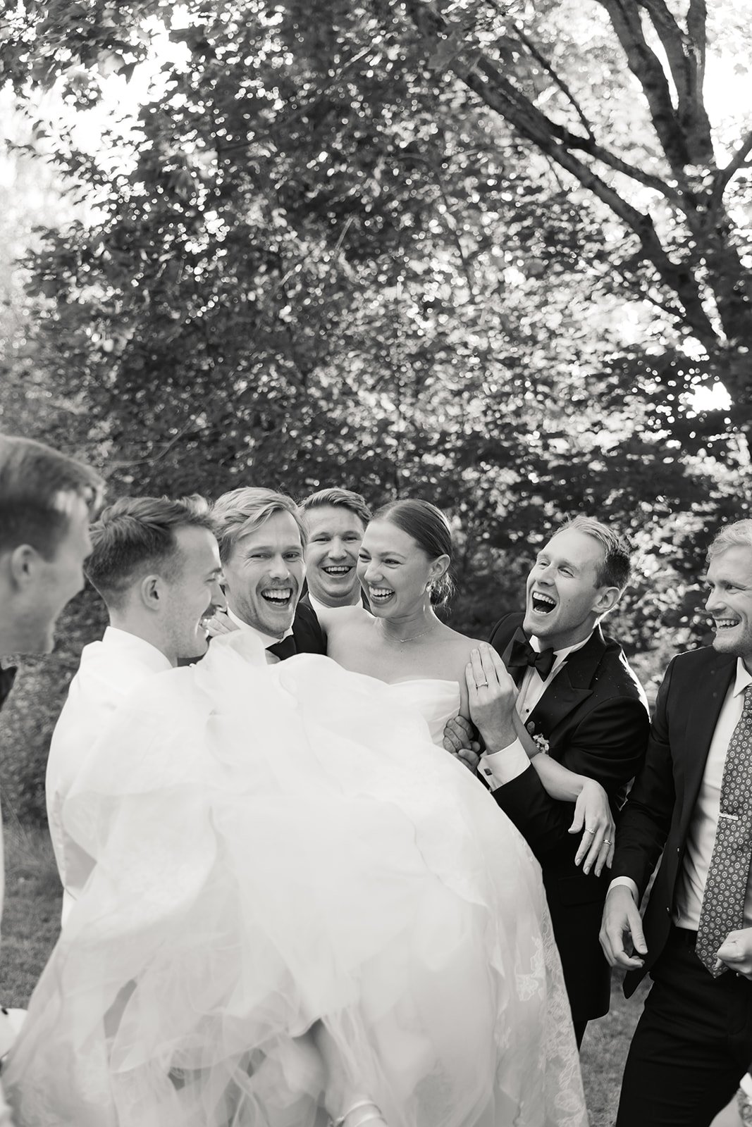 En gruppe menn i smoking og dame i brudekjole ler og holder en brud i armene ute i en skog.