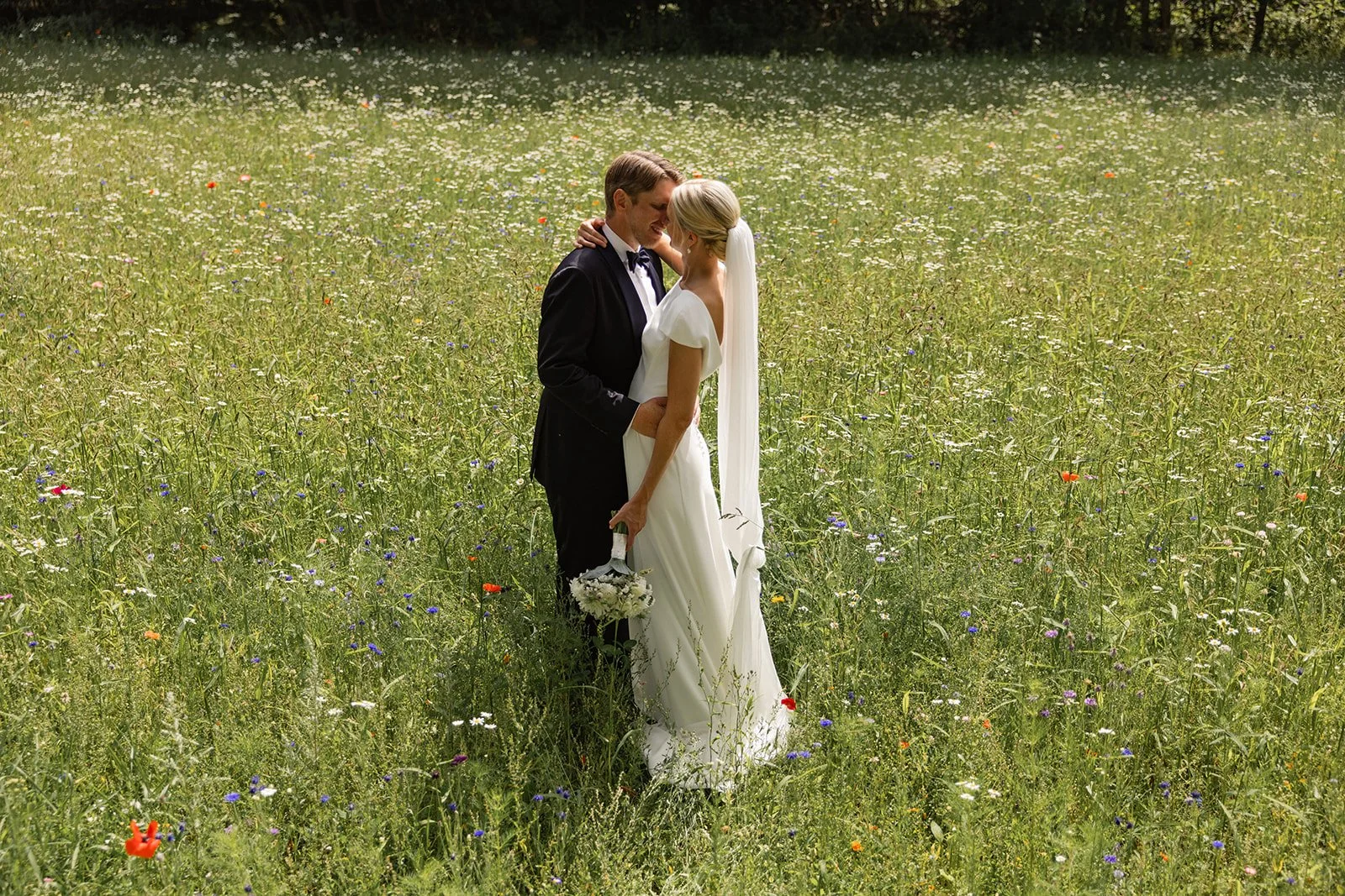 Et brudepar står i en blomstereng, de ser på hverandre, kvinnen holder en liten blomsterbukett, mannen har på seg en svart dress og sløyfe, kvinnen i en hvit kjole med slør.