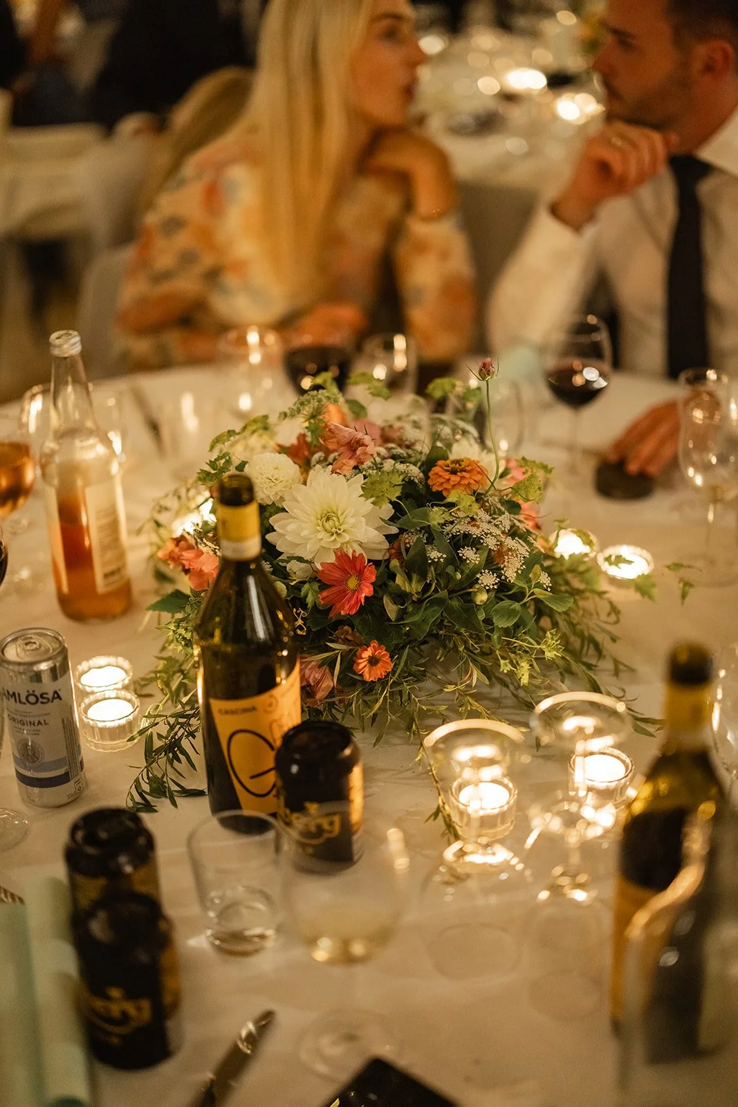 Et dekket bord med blomsteroppsats, lys, vinflasker og glass, med to personer som snakker i bakgrunnen.