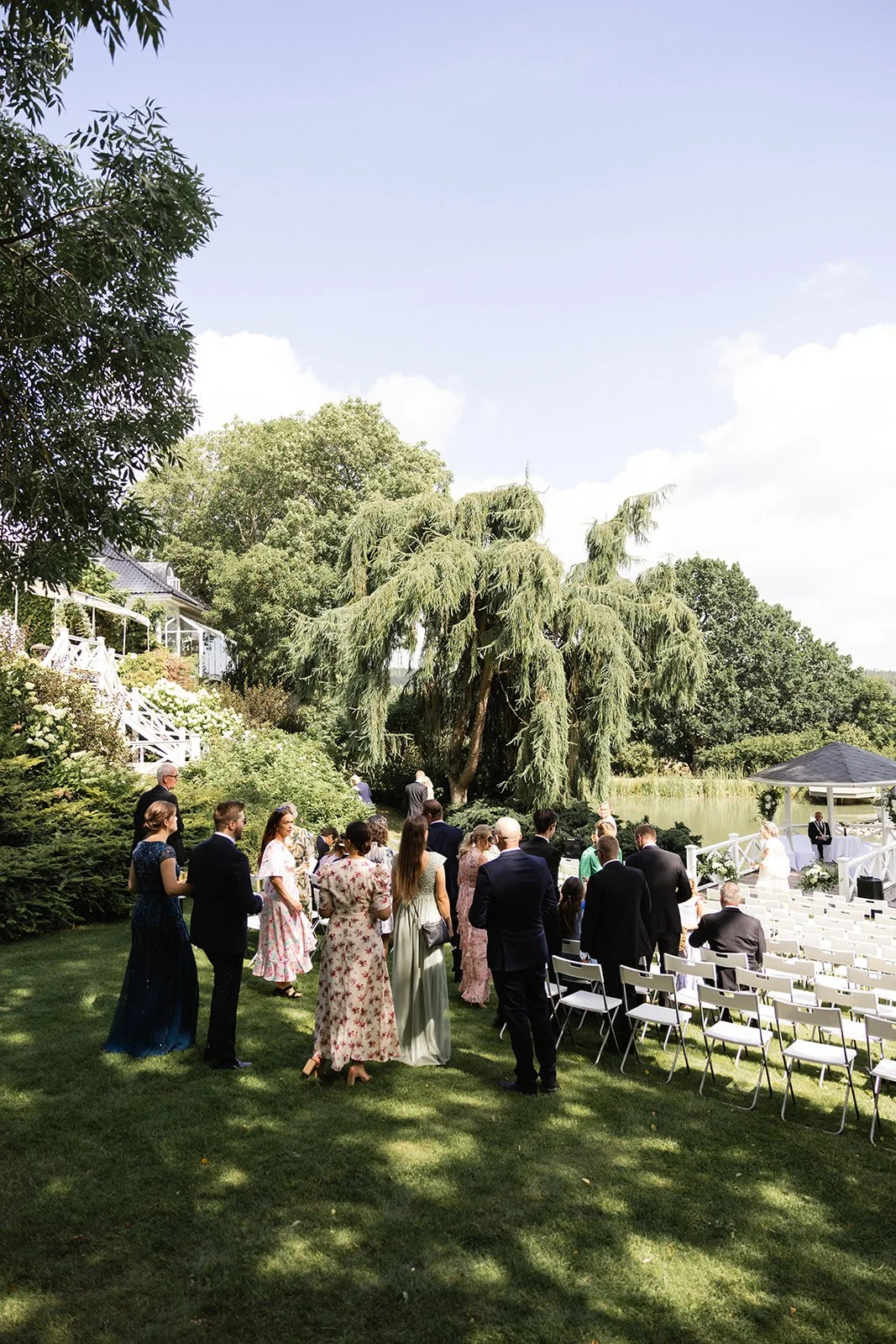 En utendørs bryllupsseremoni med mennesker i formelle klær ved en grønn hage med trær og en liten aldrende tjern. Det er solfylt og litt skyet.