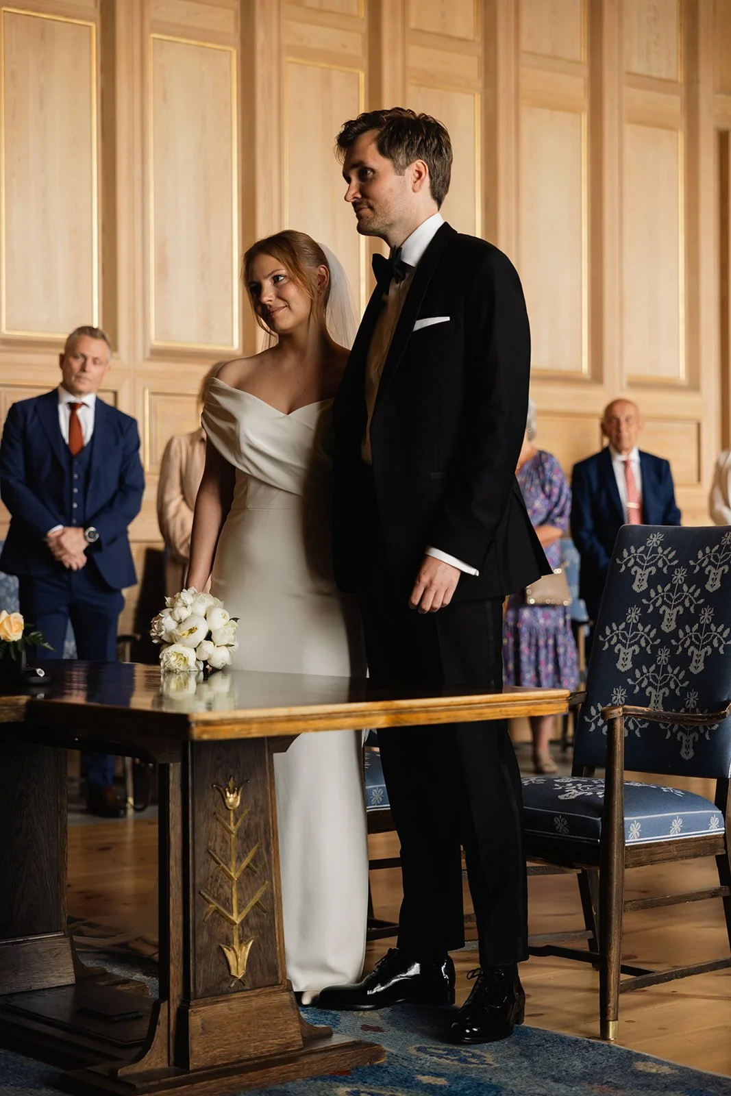 Et ektepar i bryllupsantrekk står foran en gruppe med gjester under vielsen i et rom med trepaneler. Bruden holder en bukett med hvite blomster.