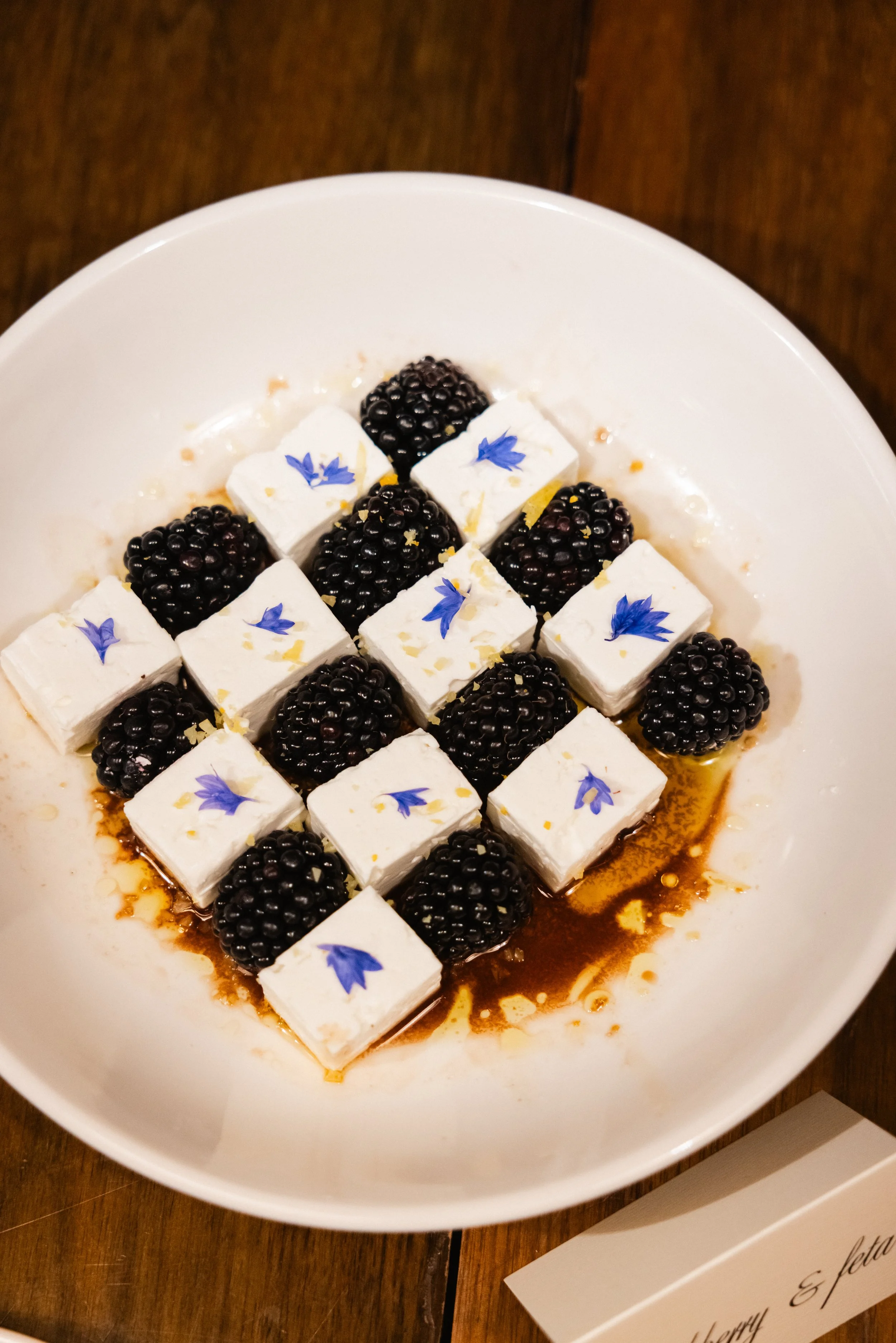 Dessert med svarte bær, små hvite mengder med krem eller marshmallow, blå blomsterdekor, brun sirup i en hvit bolle på en treplate.