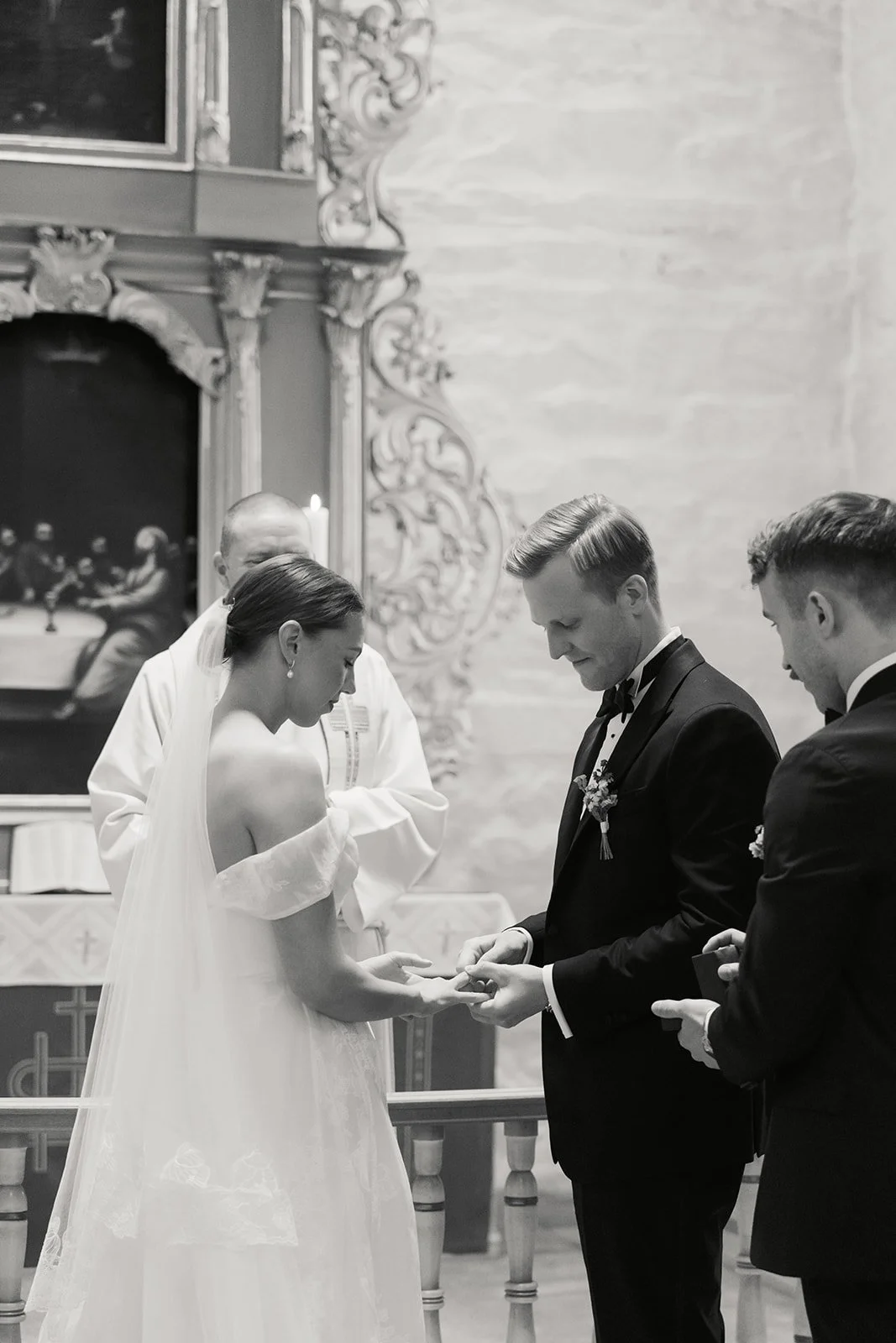 Et brudepar med brud og brudgom som bytter ringer under vielsen i en kirke, omgitt av to menn, en som driver vielsesseremonien og en tredje person.