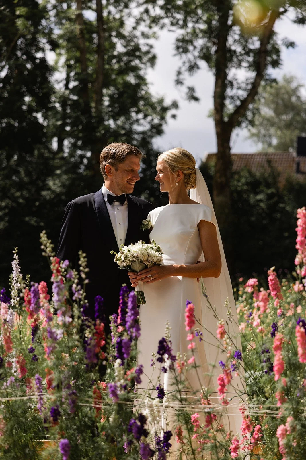 Et kledt brudepar med en mann i svart dress og en kvinne i hvit brudekjole står med blomsterbukett, omgitt av blomster og trær, under en åpen himmel.
