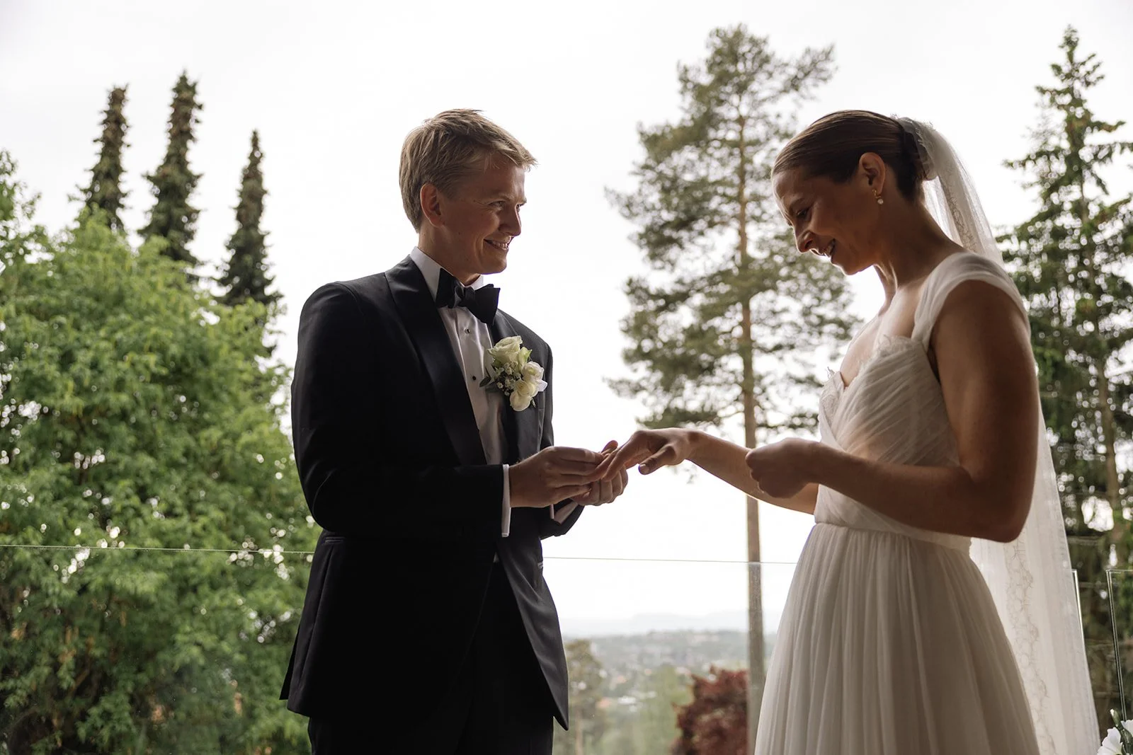 Et brudepar under vielsen, mannen i sort dress og sløyfe, kvinnen i hvit brudekjole og slør, med naturalistisk bakgrunn av trær og utsikt.
