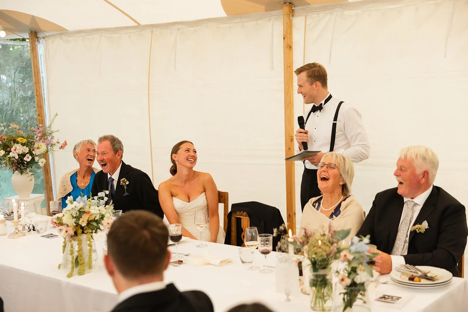 Brudeparet og familie ler og hygger seg under tale på bryllupsfest i et stort festtelt med blomsterdekorasjoner.