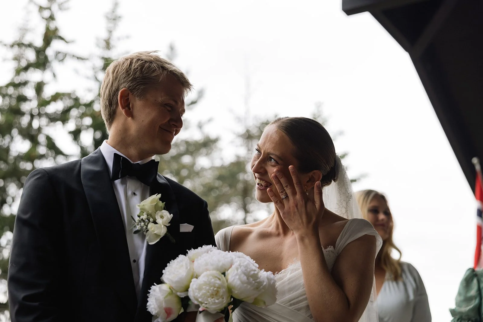 Et brudepar står sammen under en seremoni utendørs. Bruden er glad og berører ansiktet med hånden, mens hun holder en blomsterbukett. Groom er kledd i svart smoking med hvit skjorte og sort sløyfe. I bakgrunnen, en kvinne med blond hår og hvit kjole.