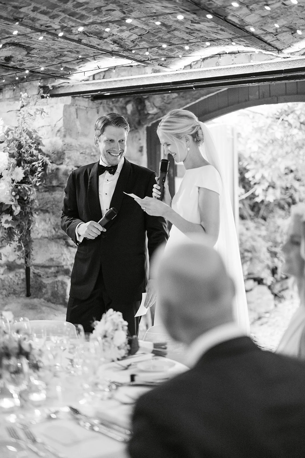 Brudepar holder en tale under et bryllup, begge smiler. Bruden leser fra et papirl apen mens hun holder en mikrofon, mens gommen smiler. Stedet ser ut til å være en innendørs setting med dekorasjoner og bord.