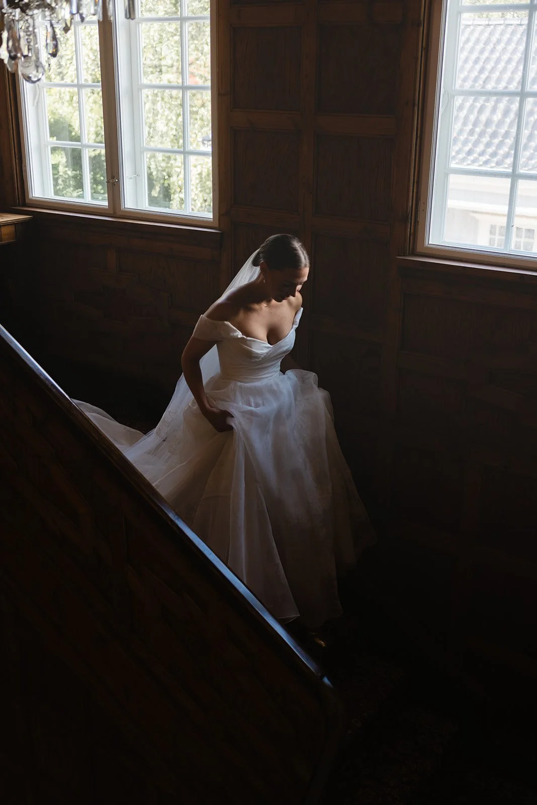 En brud i hvit brudekjole som står på en trapp innendørs ved et vindu med utsikt utendørs.
