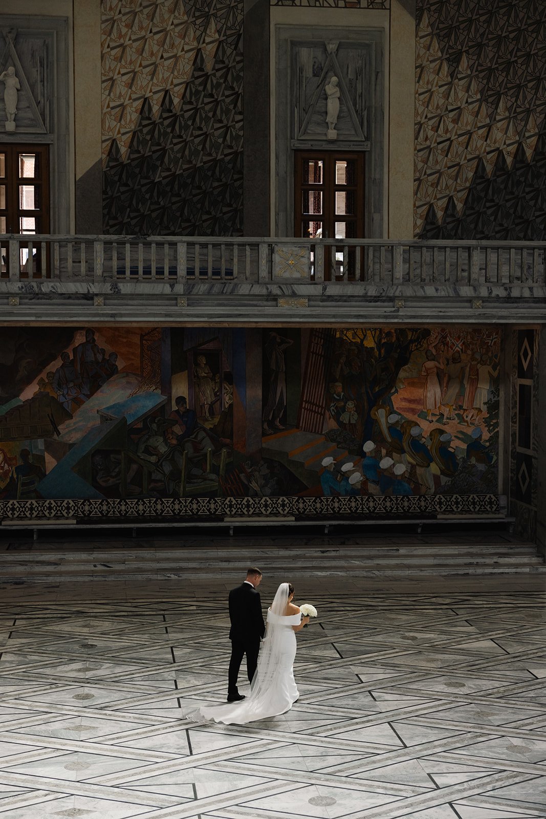 Et par i bryllupsklær står i en stor, tom hall med marmorgulv, omgitt av kunstneriske veggmalerier og arkitektoniske detaljer.