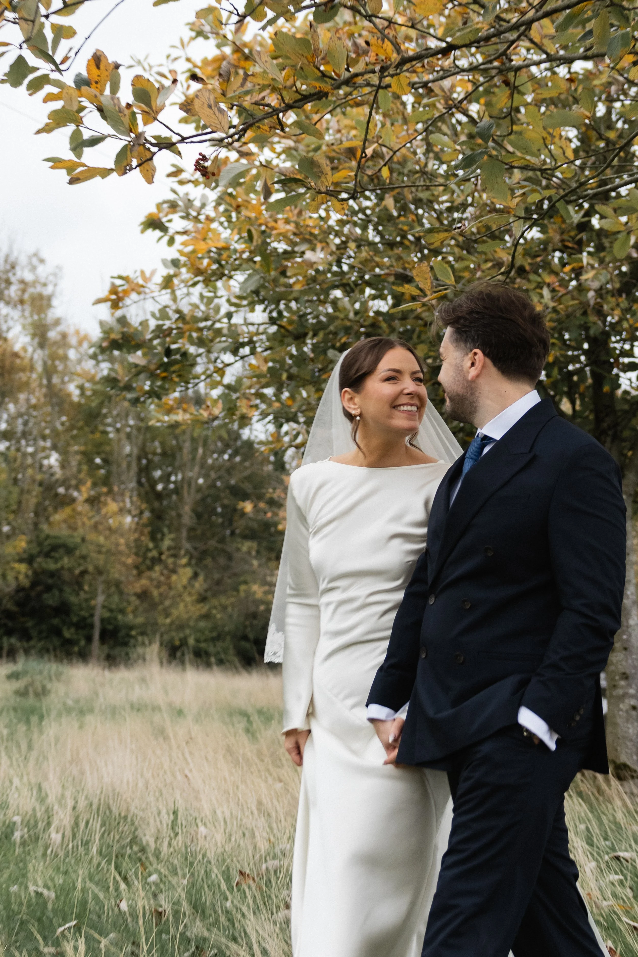 En brud og brudgom står i en åker med høstfarger i bakgrunnen, holder hender og smiler til hverandre.