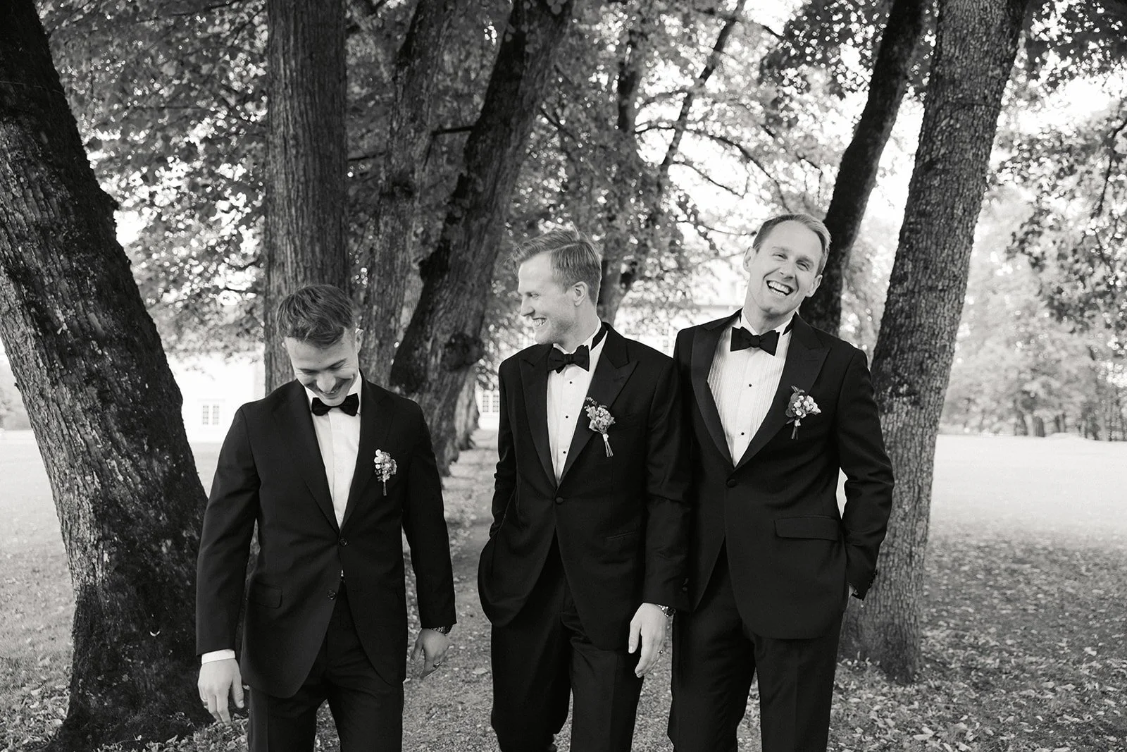 Fire menn i svarte smokinger går sammen under trær i en park, ler og har det gøy.