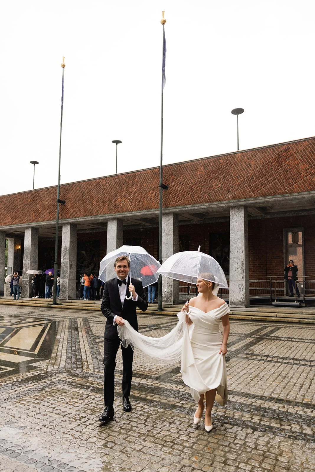 En brud og en brudgom går sammen under paraplyer i regnet utenfor en bygning med fler personer i bakgrunnen.