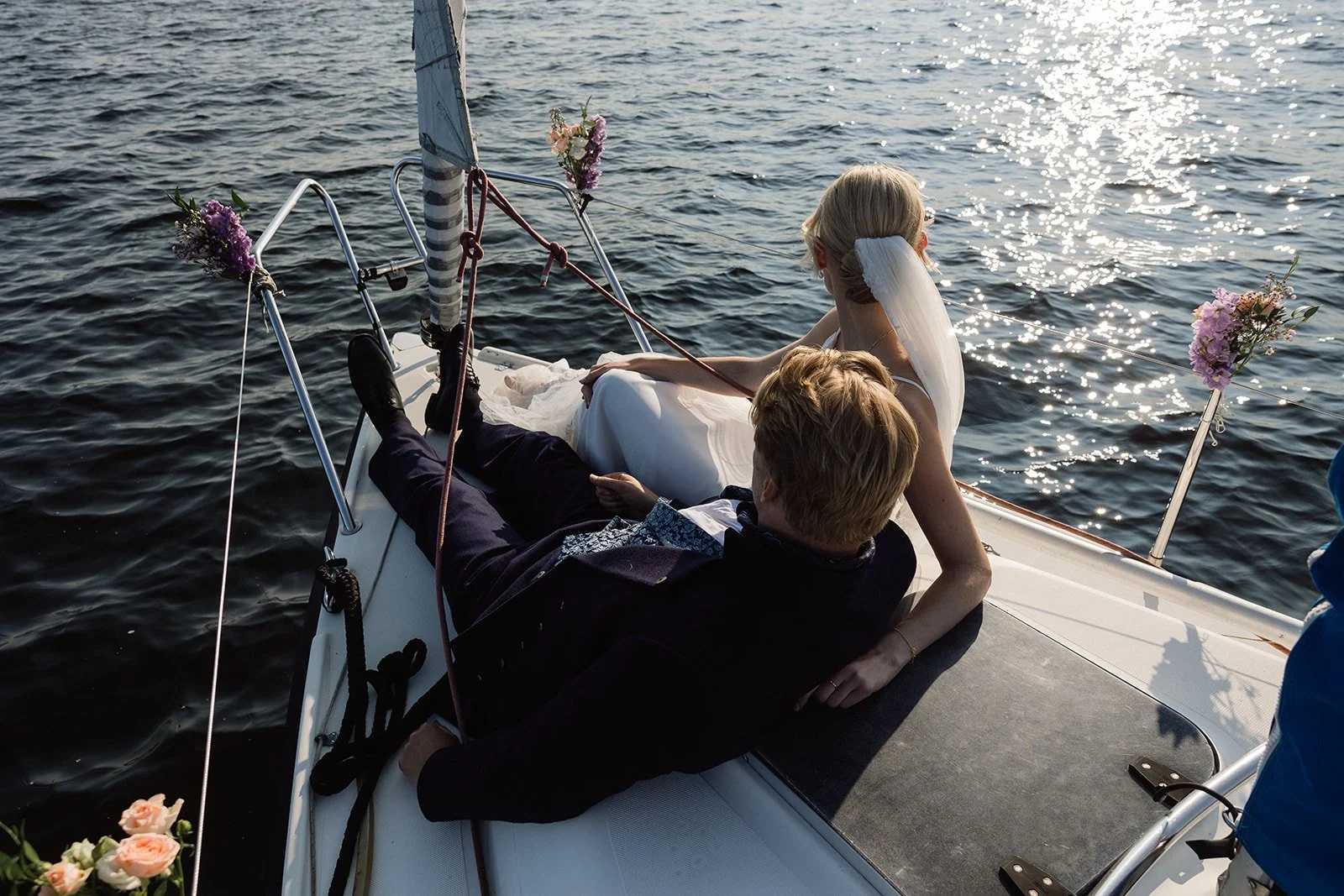 Et brudepar ligger på en båtdekk på sjøen, omgitt av blomster, i solnedgangen.