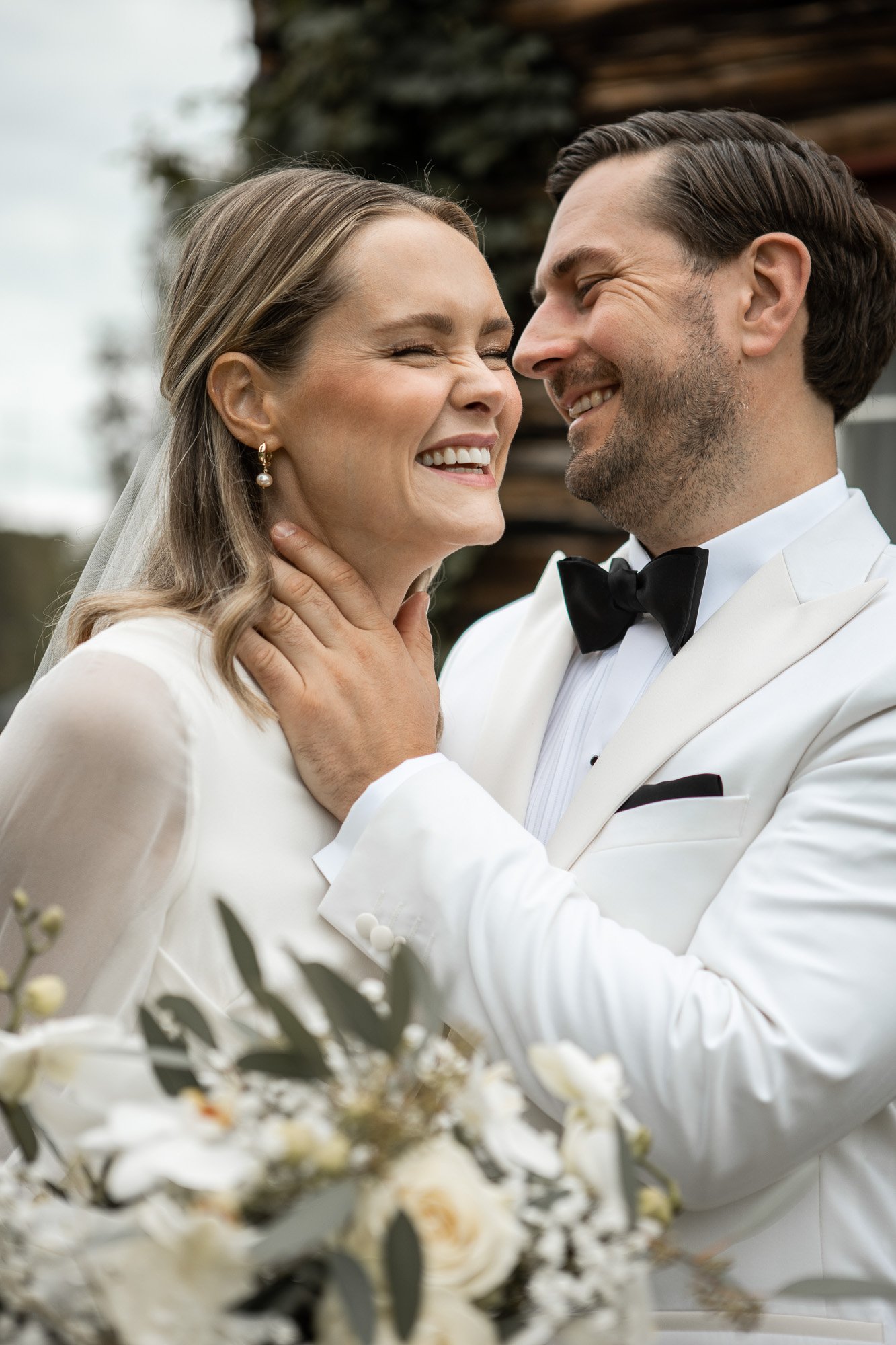 Et brudgom og en brud som smiler og byr på en kjærlig klem, kledd i elegante hvite antrekk. De står utendørs, med et tre og en bygning i bakgrunnen, omgitt av blomster