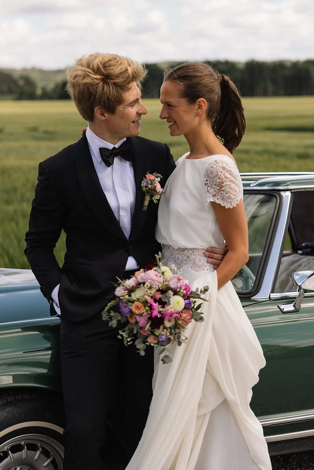 Et brudepar står foran en bil i et grønt utendørs landskap, smilende mot hverandre. Bruden holder en fargerik blomsterbukett, og begge er kledd i formell bryllupsklær.