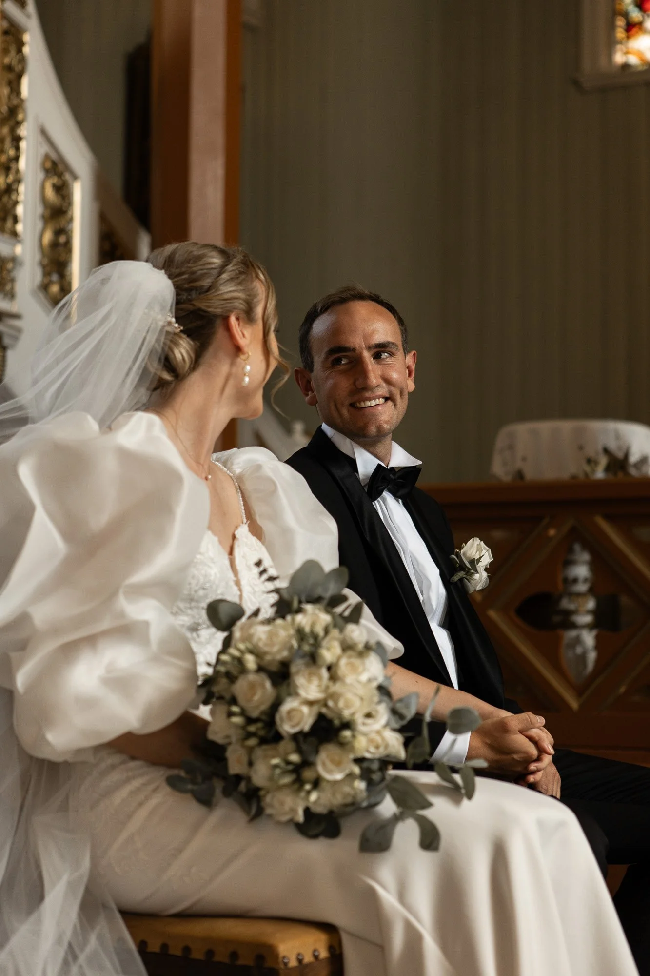 Et brudepar sitter i en kirke, kvinnen har en brudekjole og holder en blomsterbukett, mannen har smoking og slips.