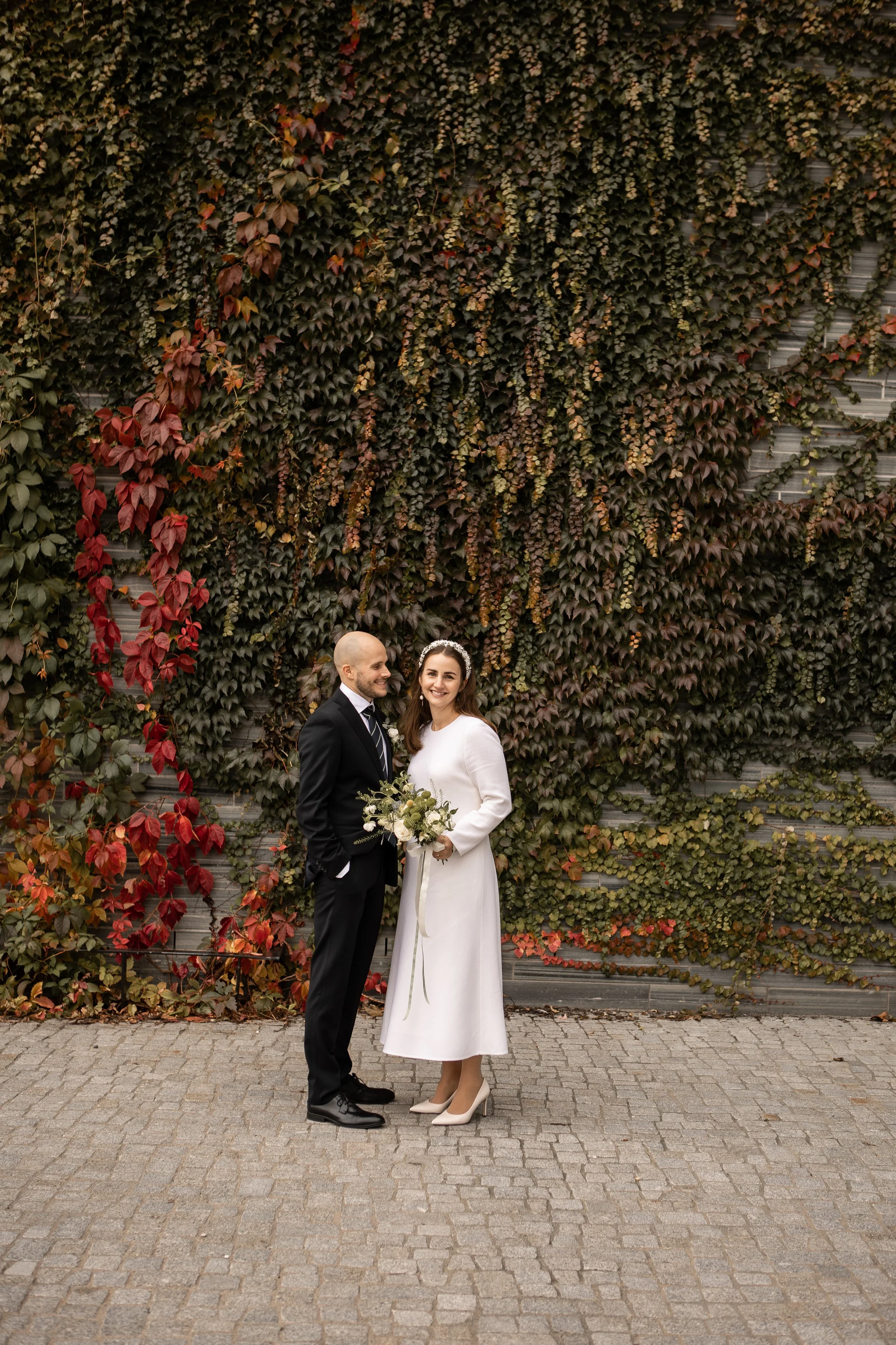 Et nygift par står foran en vegg med høstfargede blader, kvinnen har på seg en hvit kjole og holder en blomsterbukett, mens mannen har på seg en svart dress.