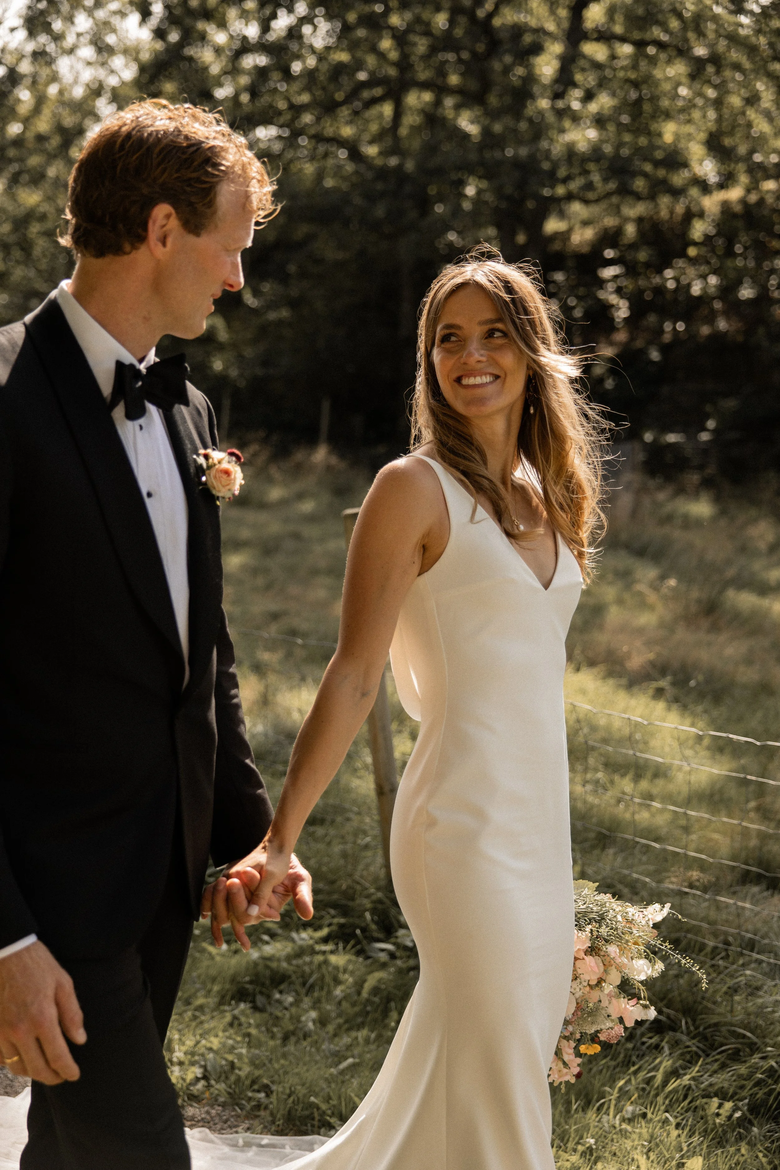 En brud og en brudgom holder hender og går sammen i et utendørs bryllup, omgitt av trær og grønt landskap, i solskinn.