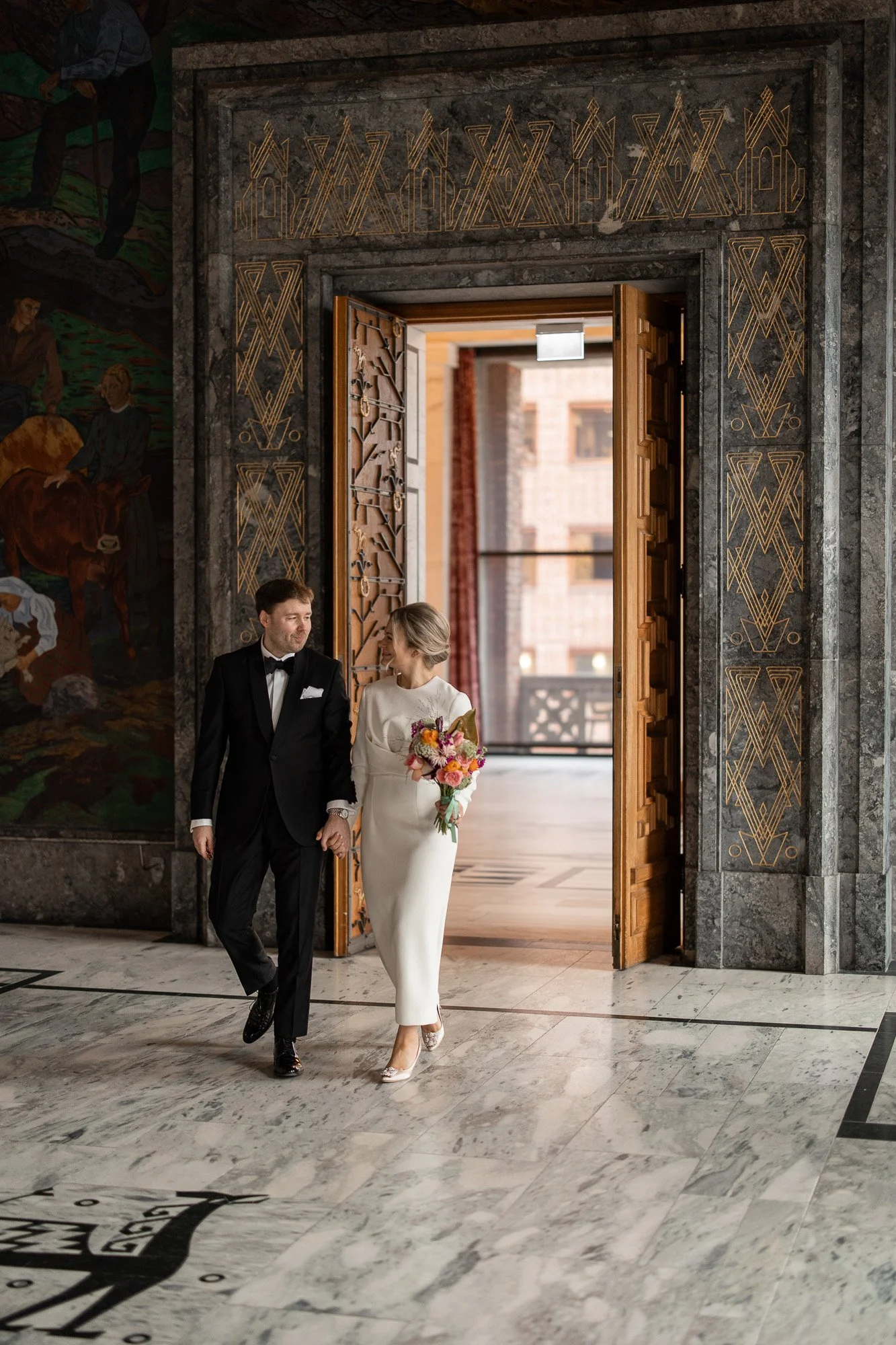 Et brudepar går inn i en stor hall med utsmykkede vegger, der bruden holder en fargerik bukett og begge smiler til hverandre.