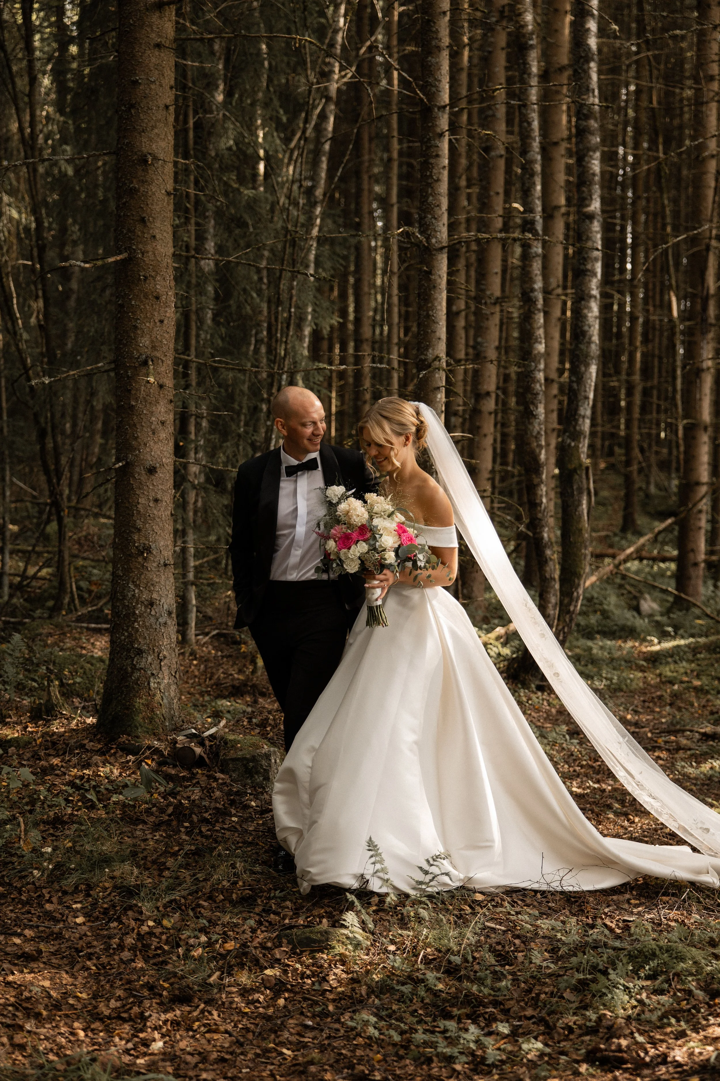 Et brudepar som går i en skog, den kvinnelige bruden holder en blomsterbukett og den mannlige brudgommen har på seg dress. Sollys skinner gjennom trærne.