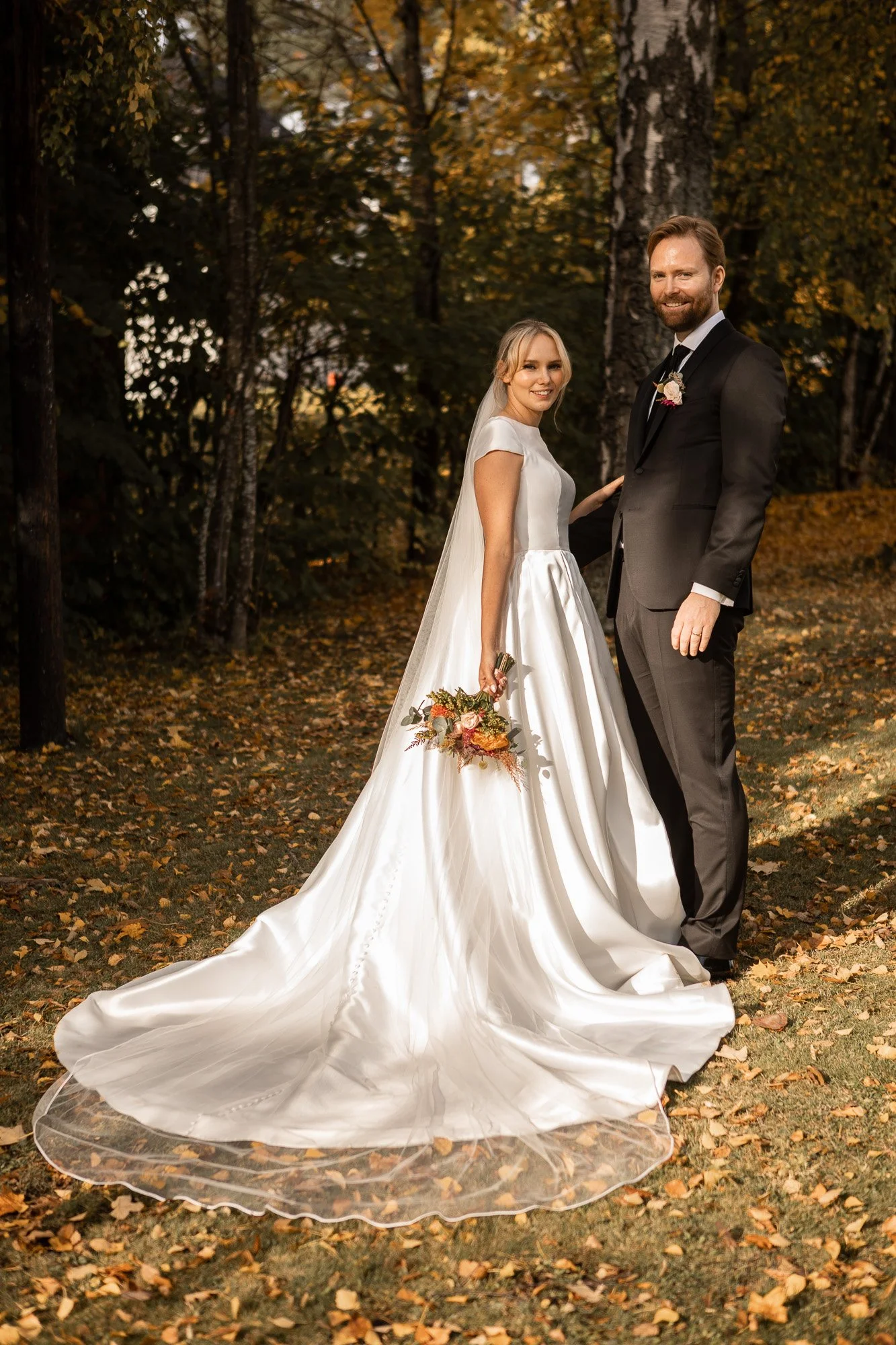 En brudepar står i en skog om høsten. Kvinnen bærer en hvit brudekjole og holder en blomsterbukett, mens mannen har på seg smoking og har en boutonnière. De smiler til kameraet.