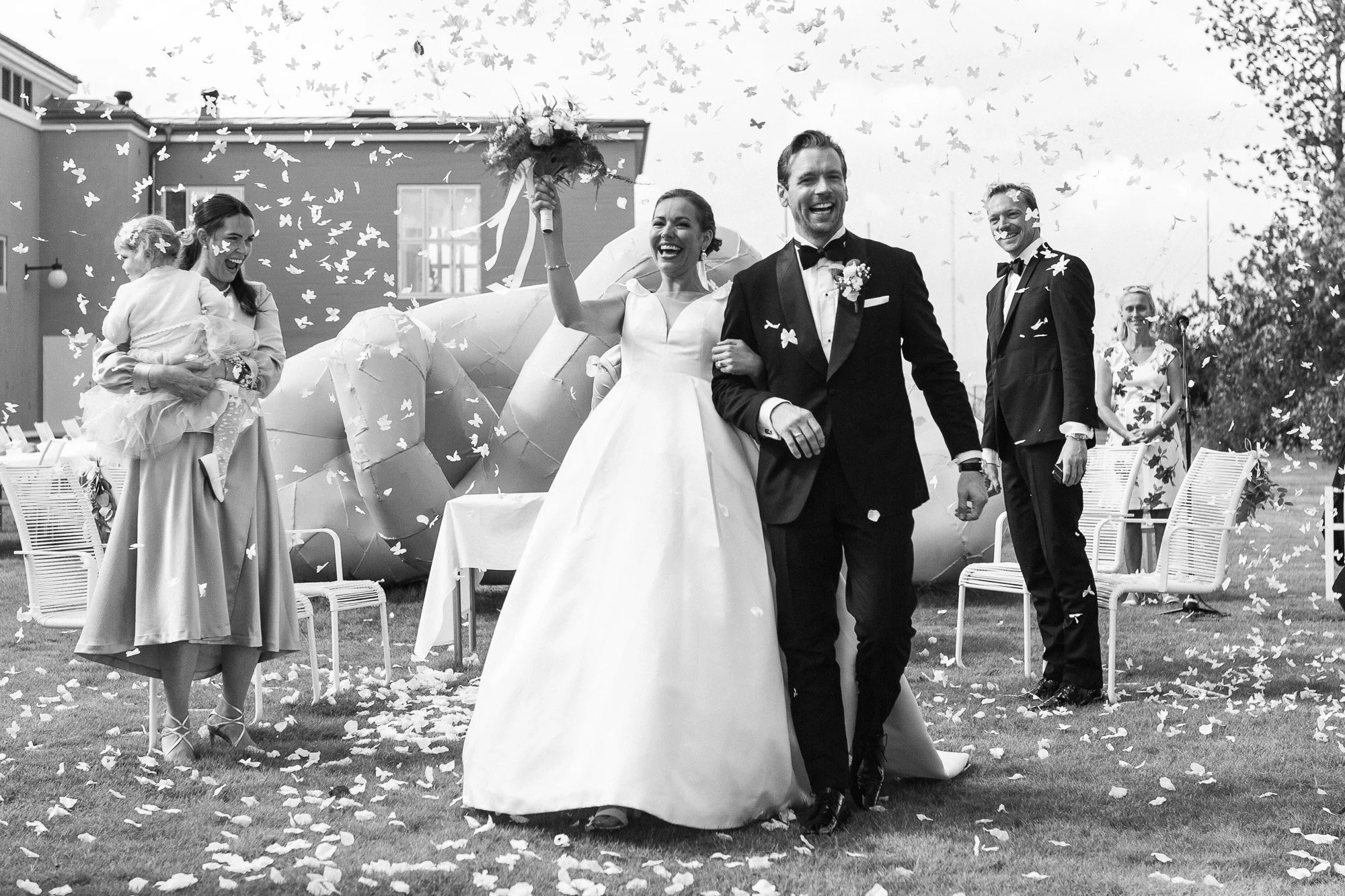 Bryllupsceremoni med den lykkelige bruden og brudgommen som går ut. Glad brud med blomsterflor og brudgommen i dress, omgitt av fornøyde gjester som kaster blomster blant blomsterblad på bakken.