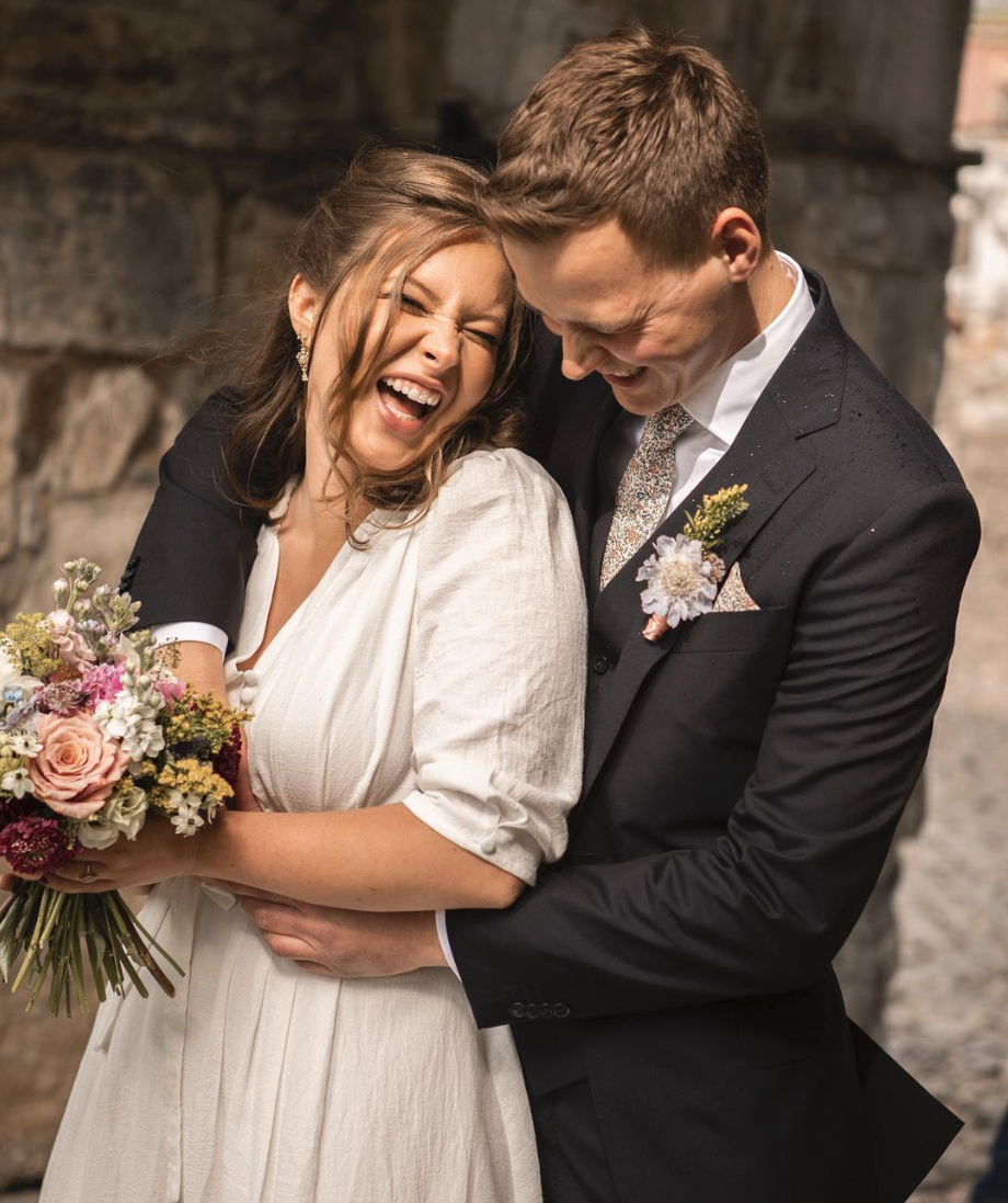 Et lykkelig ektepar på bryllupsdagen, med mannen i dress og kvinnen i hvit kjole, som ler og holder en blomsterbukett.