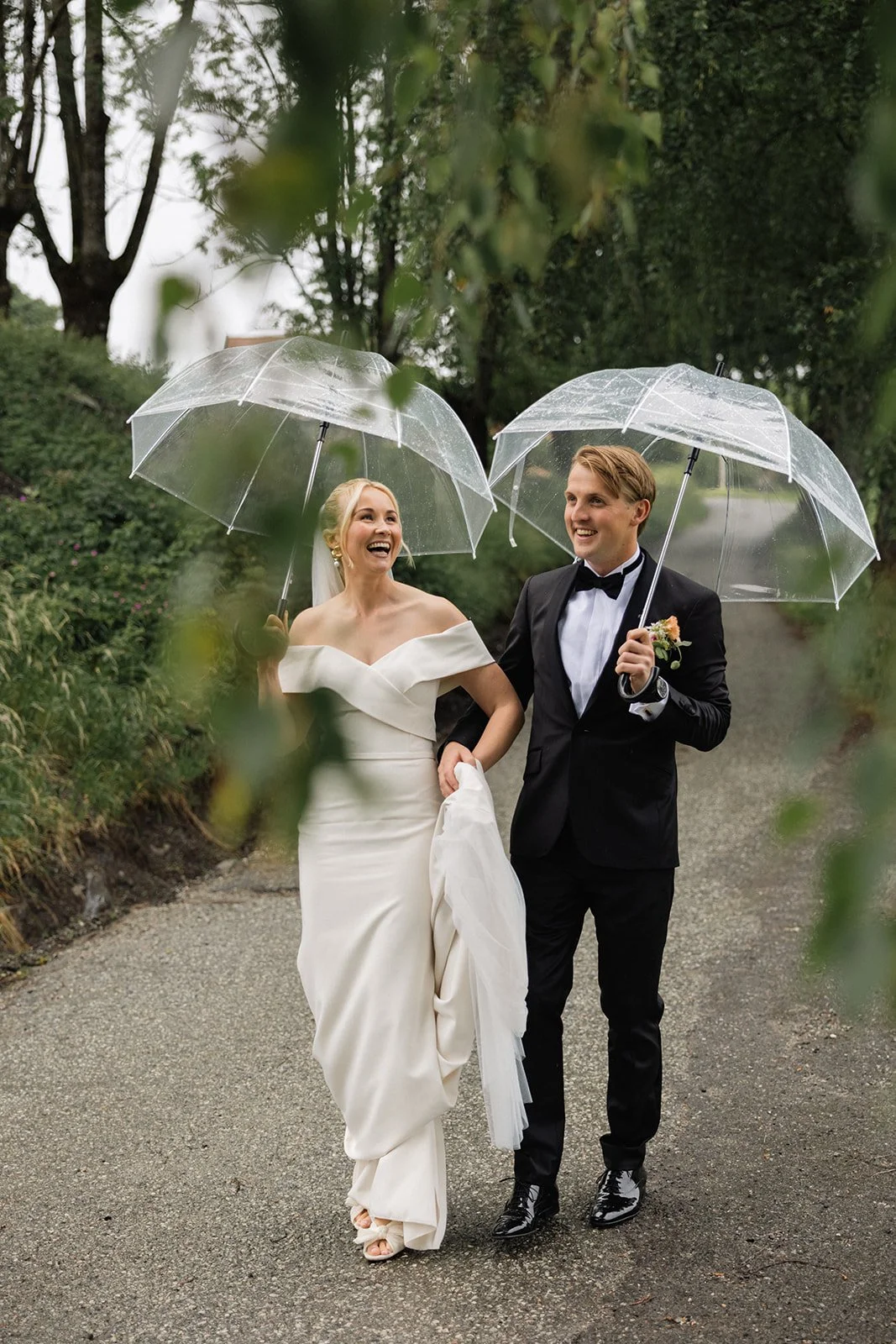 En brud og brudgom går hånd i hånd under regn med klare paraplyer i en grønn skogssti, smilende og glade.