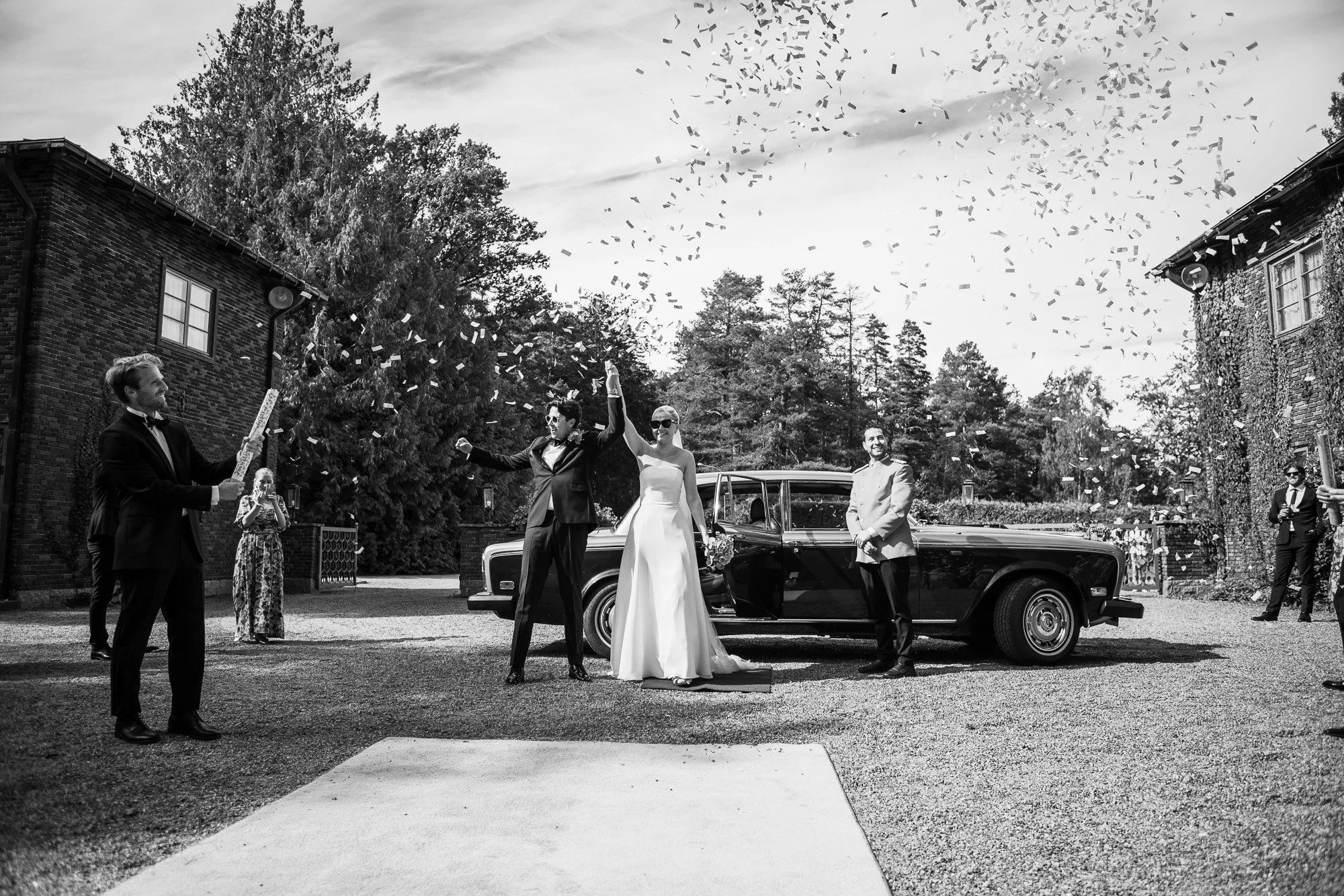 Sort-hvitt bilde av et bryllup utendørs med bruden og brudgommen som feirer, omgitt av venner og familie, med konfetti i luften, en bil parkert bak, trær og bygninger i bakgrunnen.