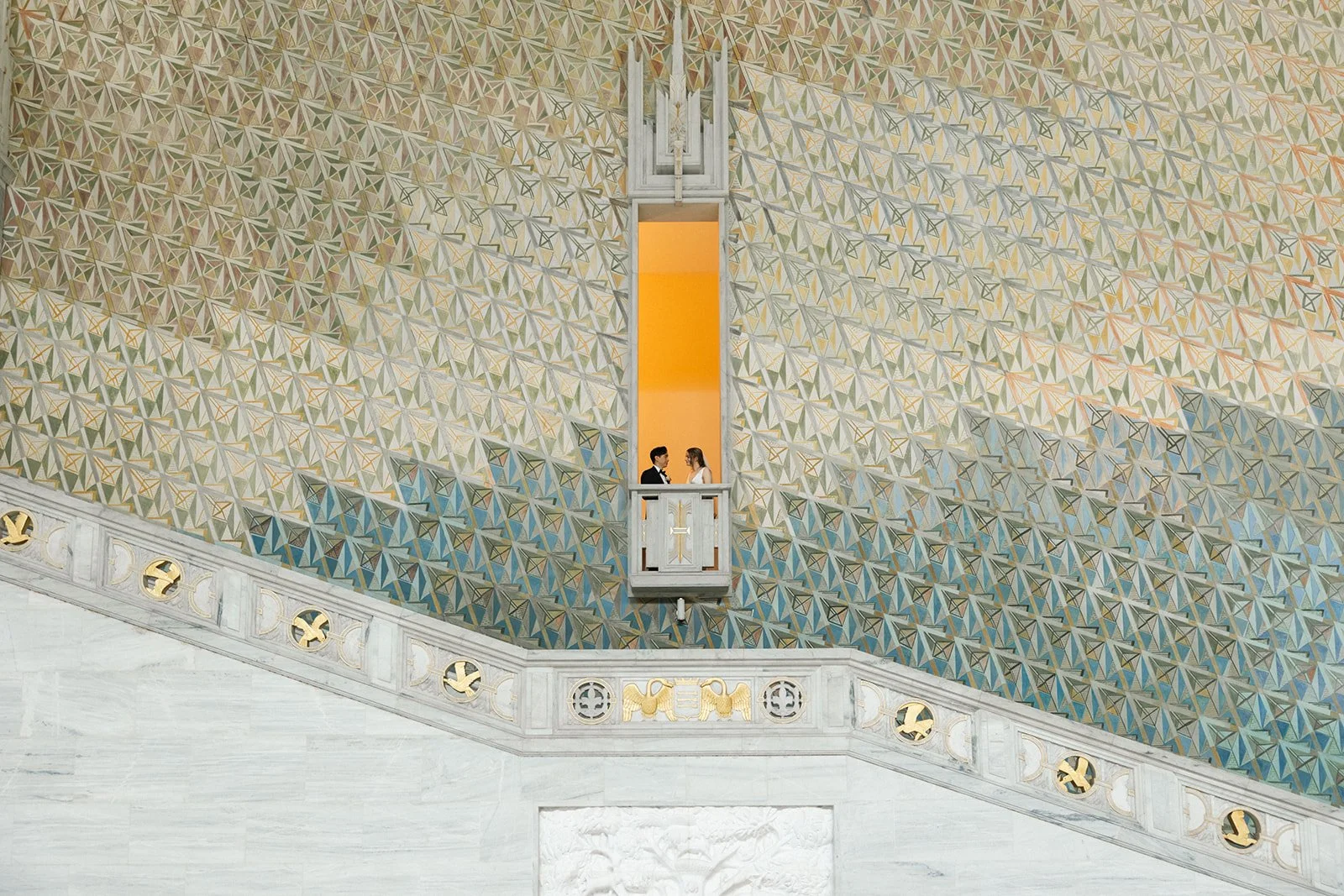 To personer står på en liten balkong i et stort rom med utsmykket vegg, med geometrisk mønster i gull, grønt, blått og beige. Oslo Rådhus.