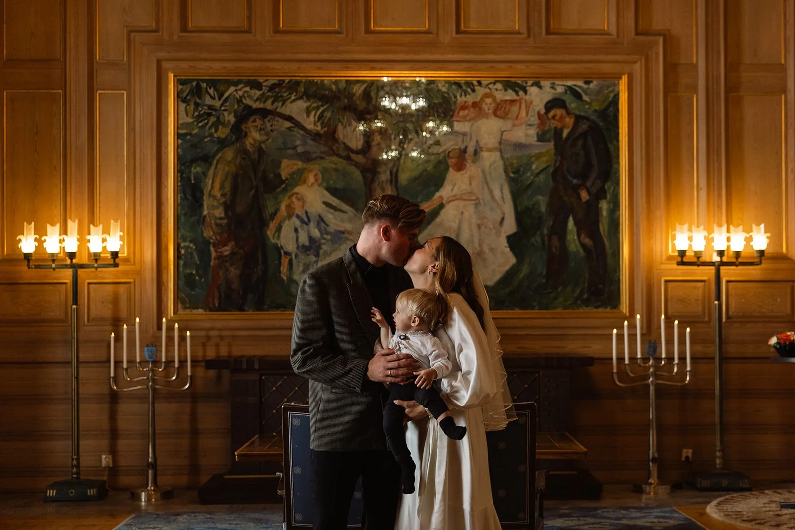 Et ungt par i formelle klær og et lite barn kysser hverandre i et rom med treveggpaneler, store lysestaker og et stort maleri i bakgrunnen.