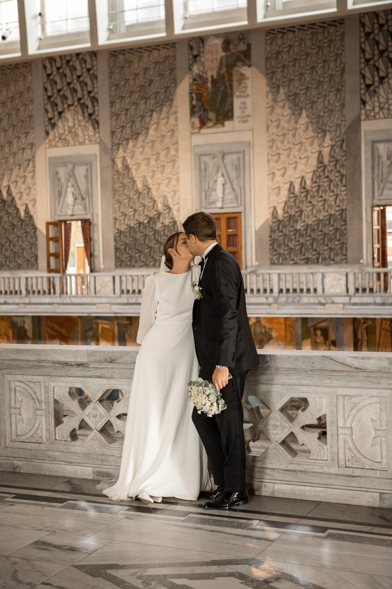 Et brullupsbilde av et ungt par som kysser, med kvinnen i en hvit brudekjole og mannen i en svart tuxedo, inni en stor og kunstnerisk kirke.