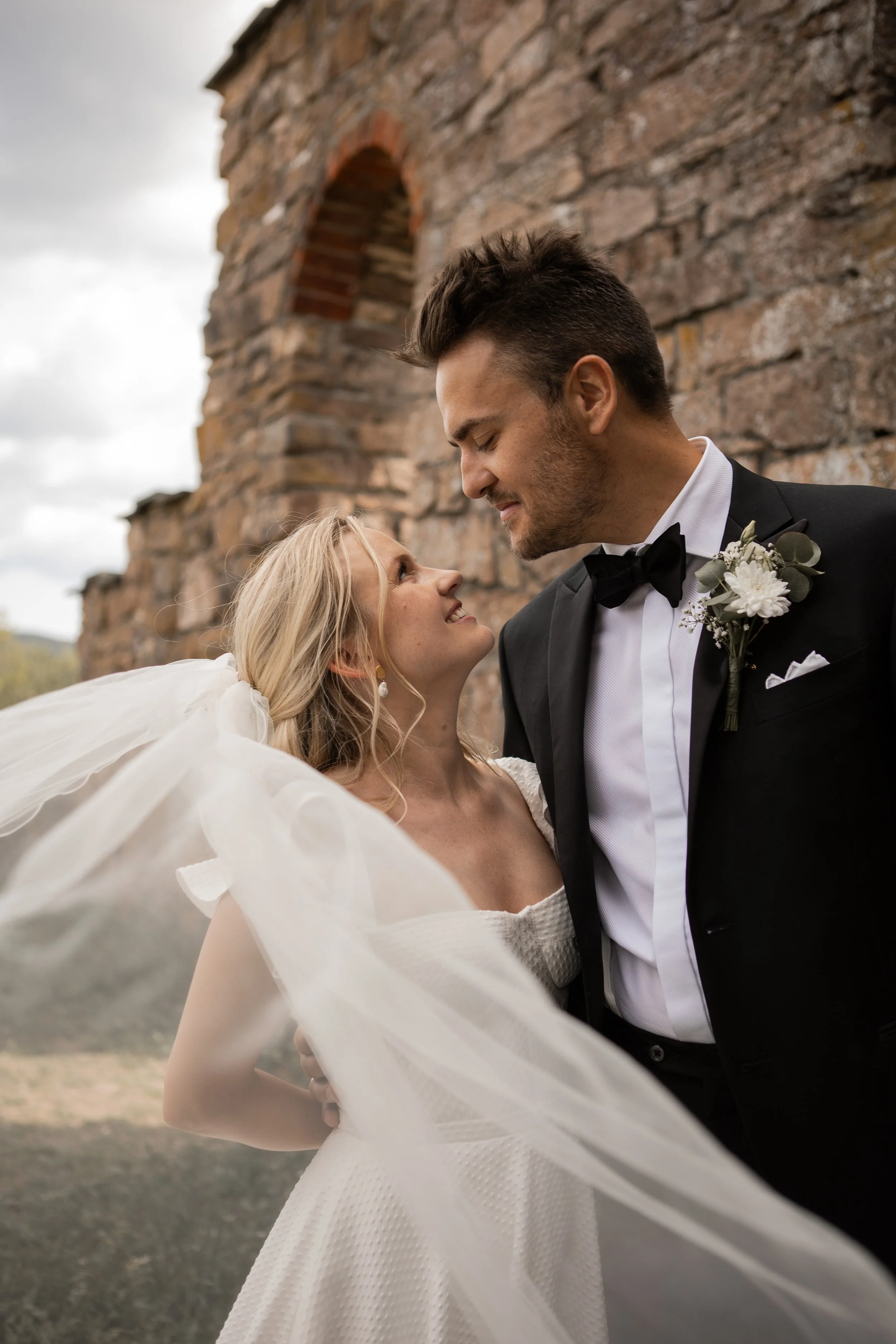 Et brudepar står nær hverandre med ansiktene tett, foran en gammel steinmur, i en kjærlighetsfull og intim øyeblikk.