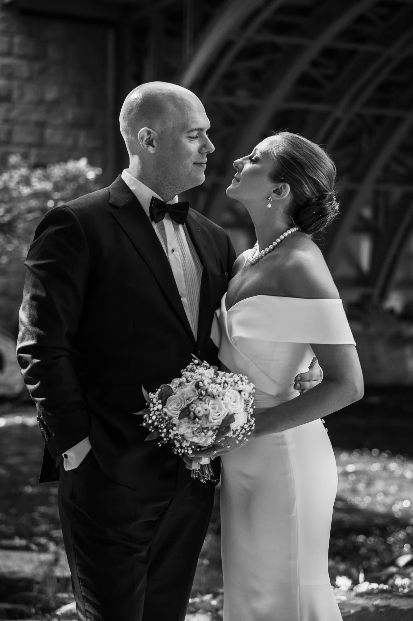 Et sort-hvitt bilde av et brudepar som står tett sammen, solen skinner på dem, med en bro i bakgrunnen.