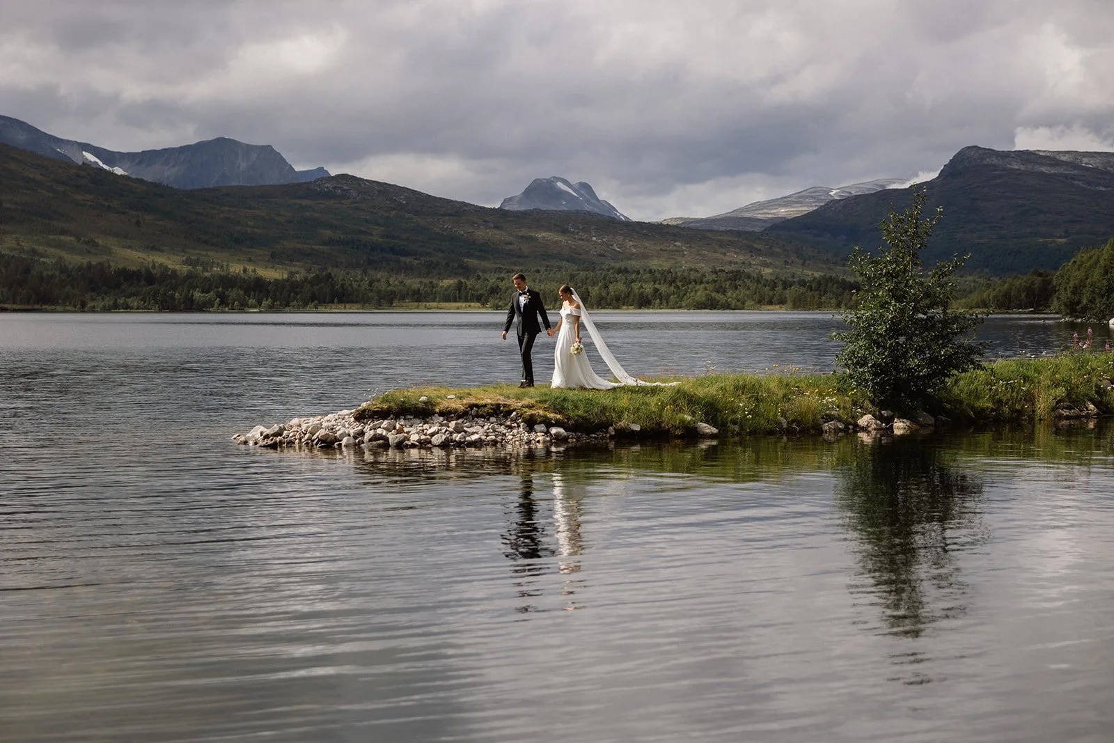 En nygift brud og brudgom som går hånd i hånd på en liten landtunge i en innsjø, omgitt av fjell og skog, med overskyet himmel.