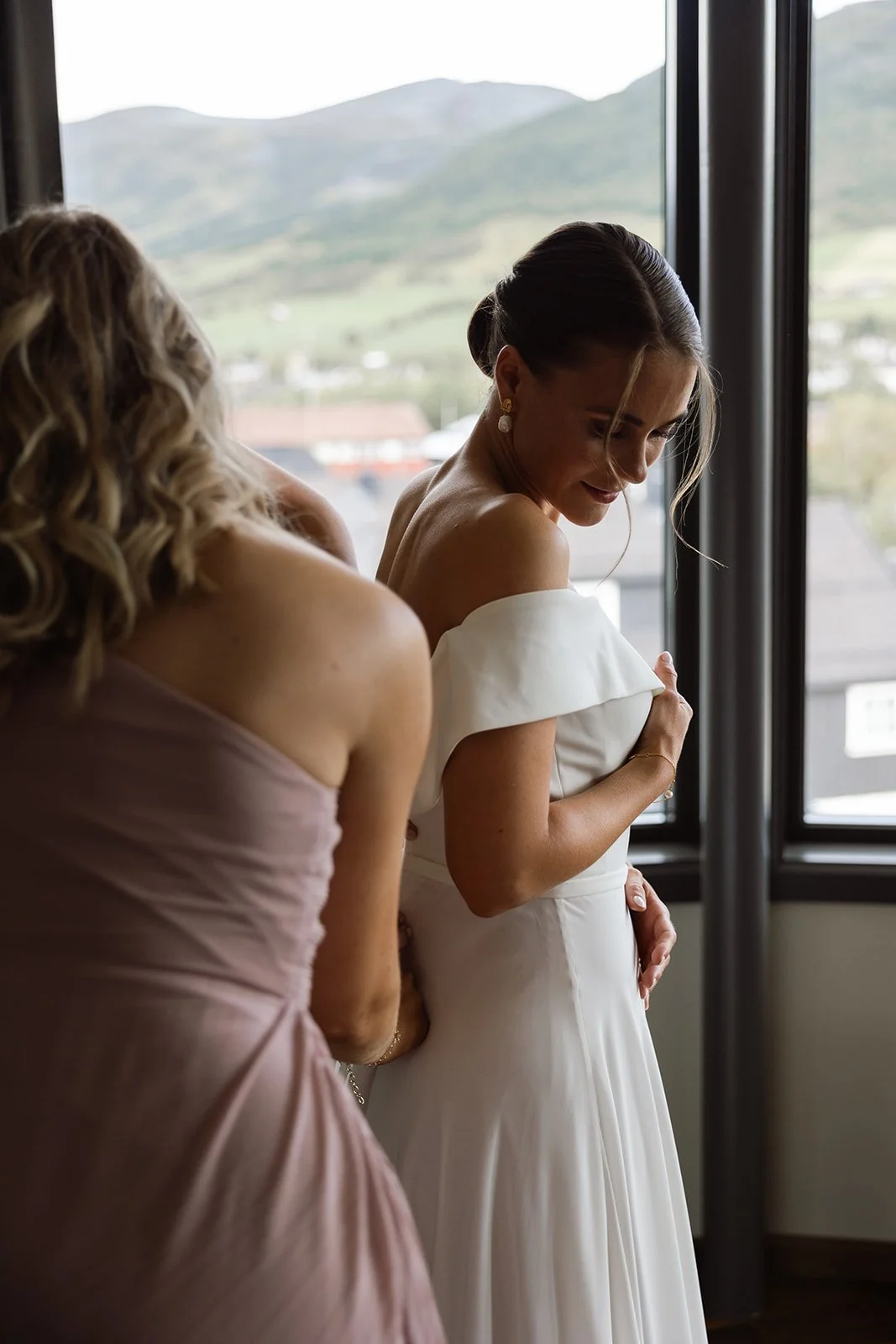 To kvinner står ved et vindu, en i en hvit kjole og den andre i en rosa kjole, og nyter øyeblikket.
