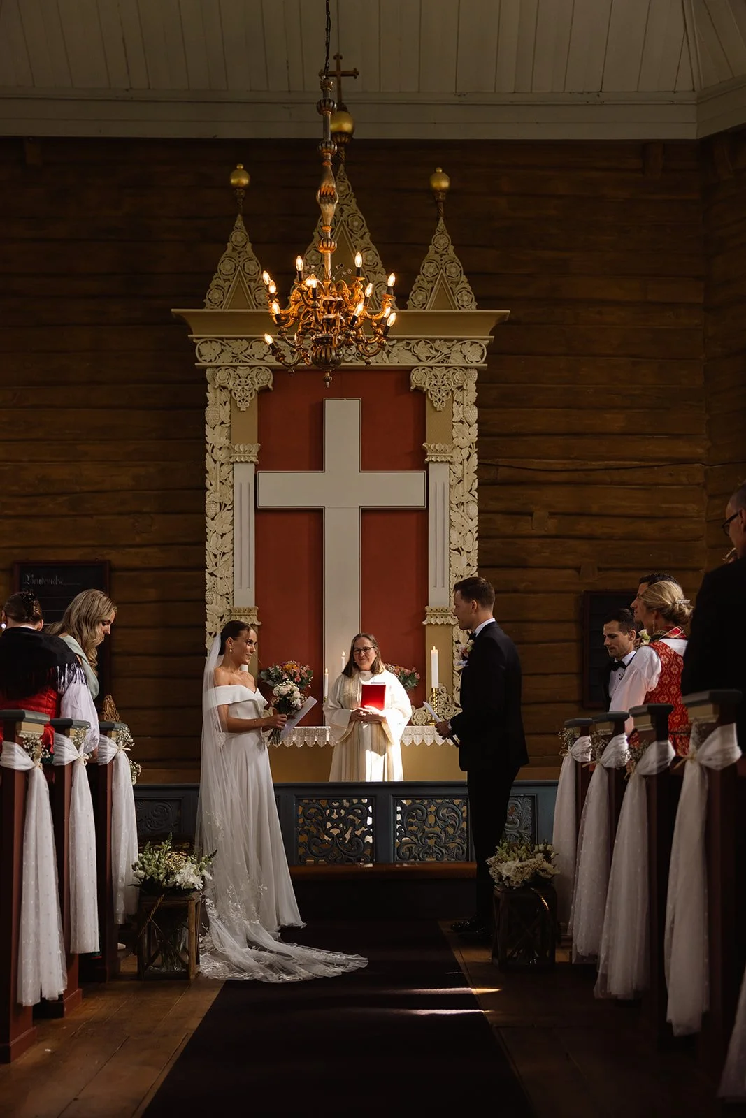 Et bryllup i en kirke med bruden og brudgommen foran alteret, omgitt av vielsesvitner og prestinne. Bruden holder en bukett, og prestinnen har en bok og et lys. Det er et kors bak alteret, og kirken har tre pærer på en lysekrone over.