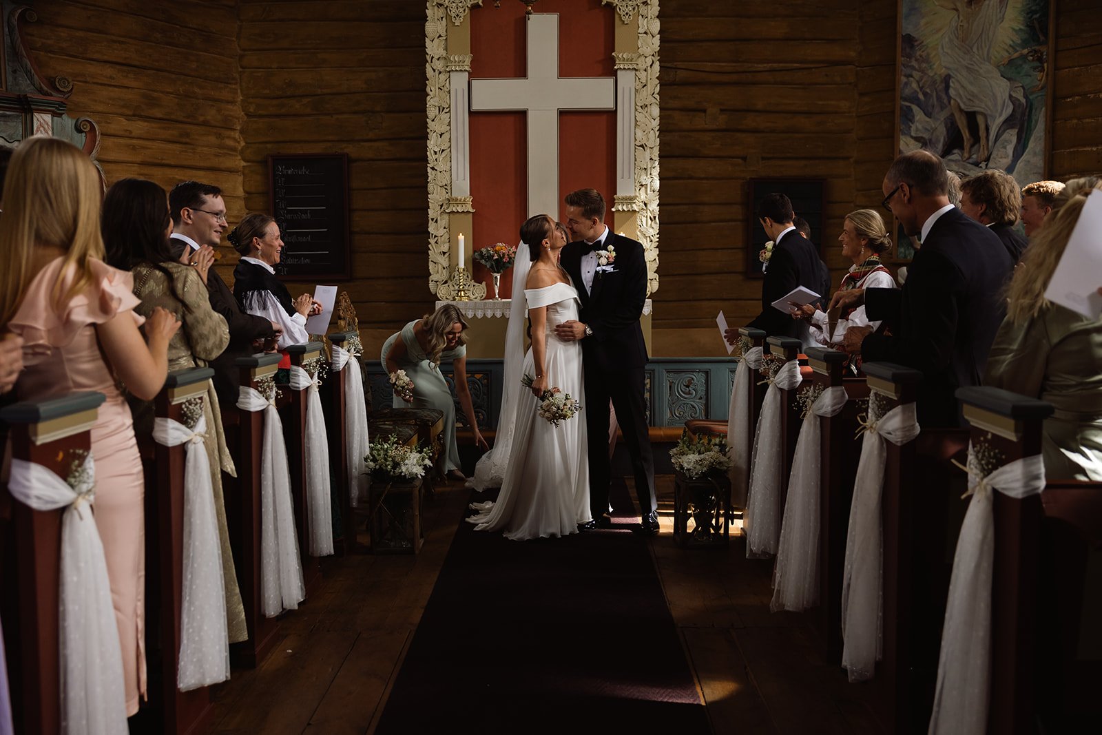 Et brudepar kysser under vielsen i en kirke med familien og venner som observerer.