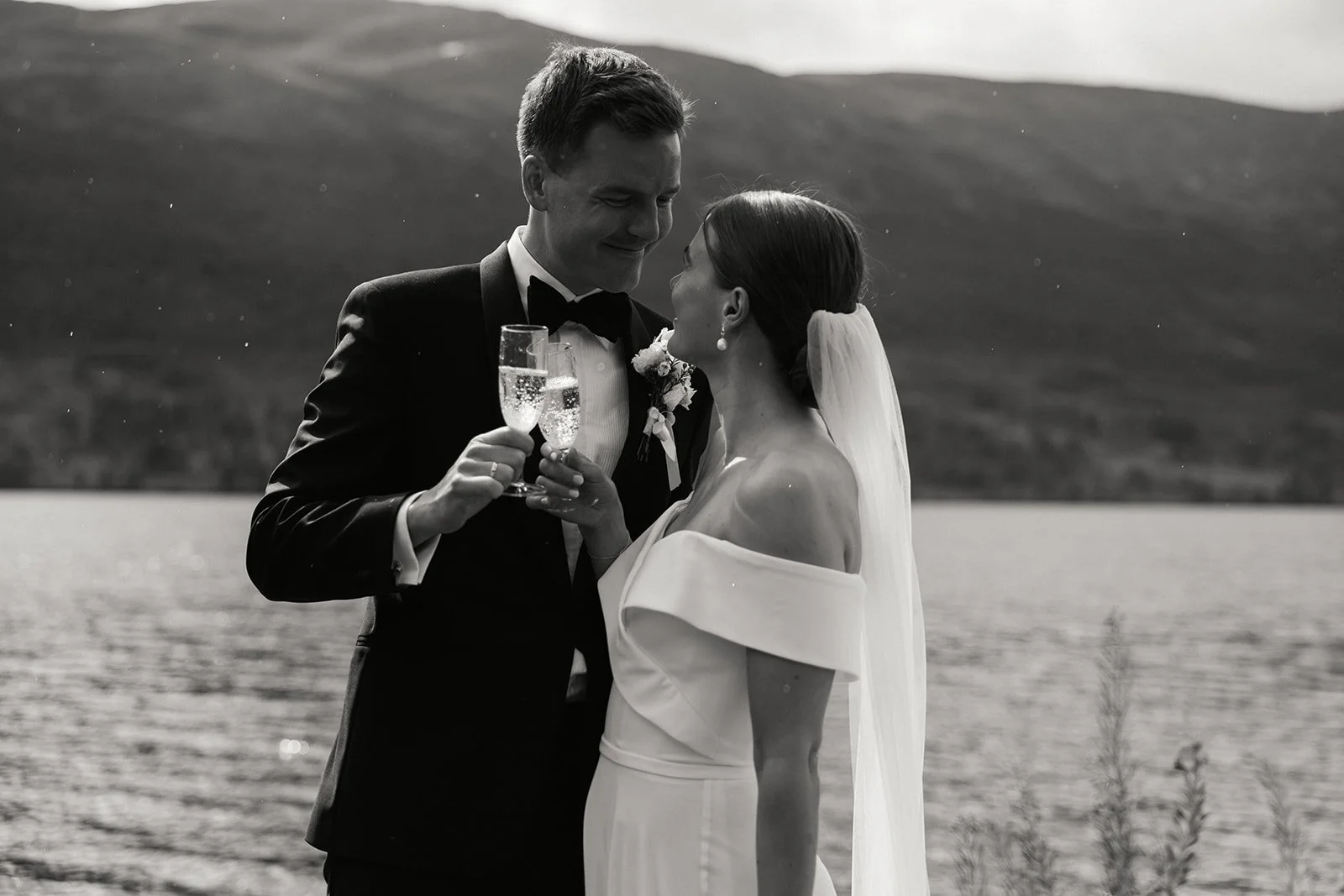 Et bryllupsbilde av en brud og brudgom som feirer med champagne ved en innsjø med fjell i bakgrunnen, i svart-hvitt.