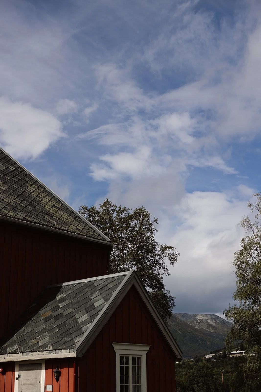 Bilde av en liten rød hus med hvite vinduer og tak av grå takstein, omgitt av trær og fjell i bakgrunnen under en skyet himmel.
