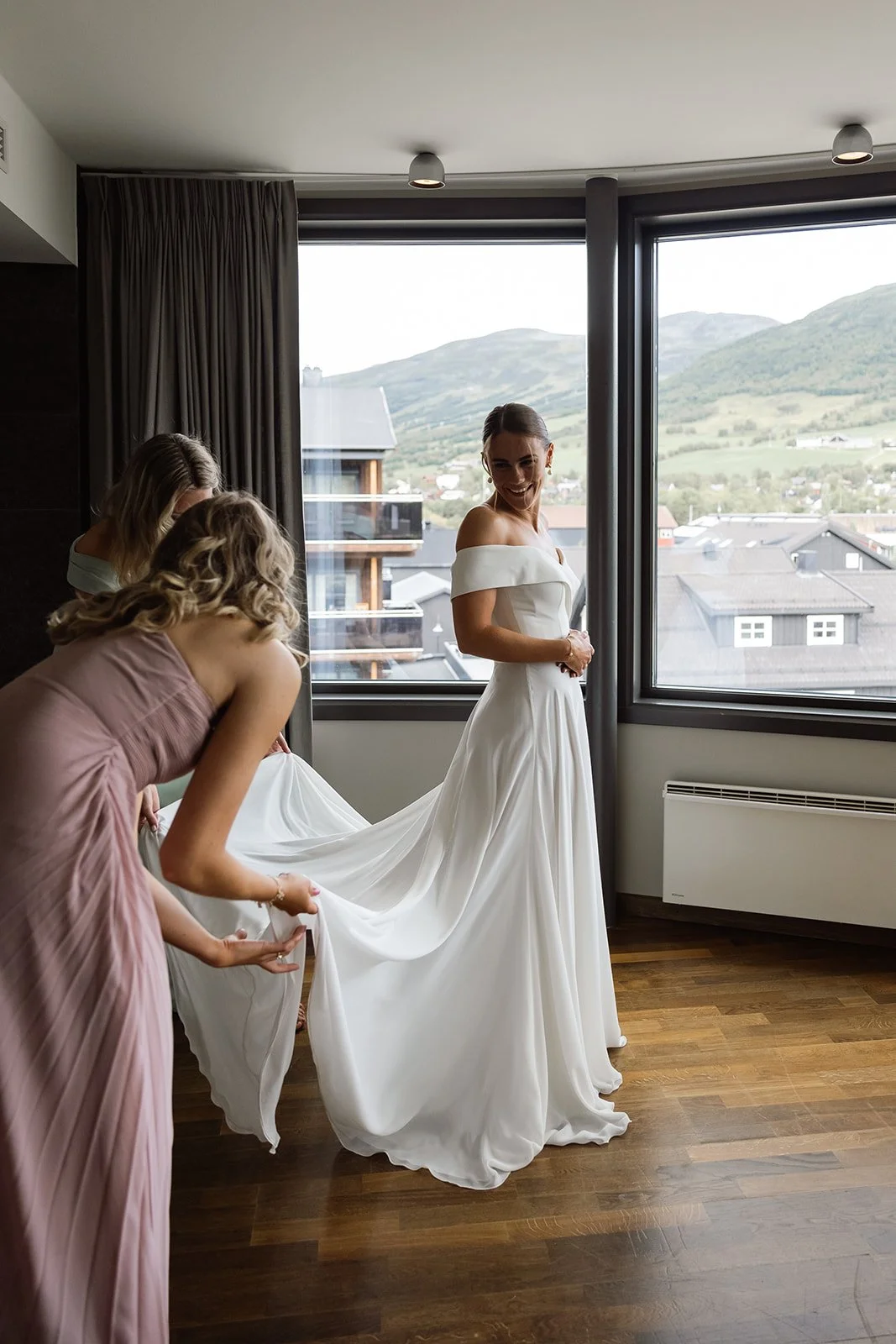 En brud i en hvit bryllupskjole får hjelp med kjolen av to kvinner i et rom med store vinduer og fjellutsikt.