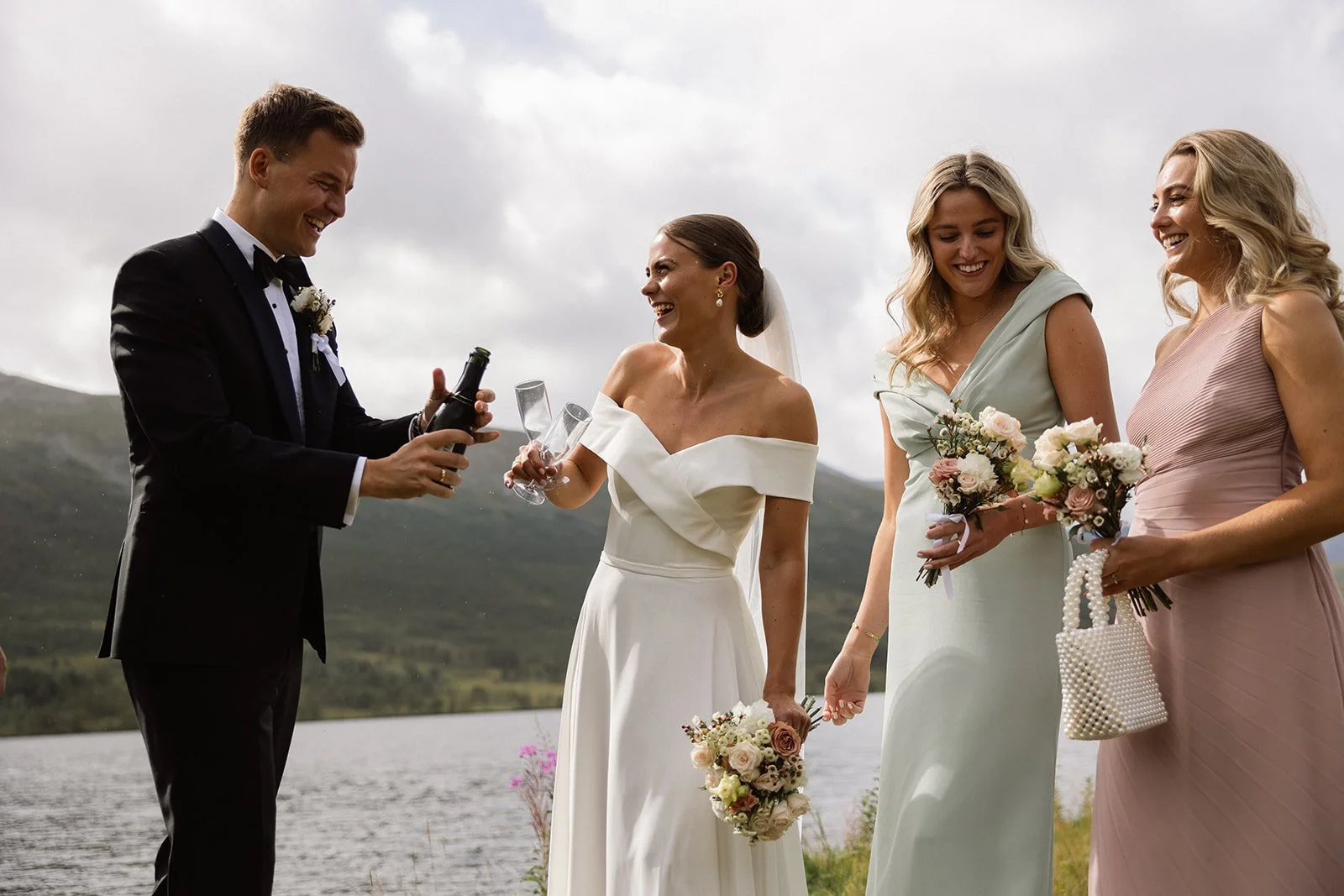 En gruppe mennesker i brudeklær blir feiret ved et vann med fjell i bakgrunnen.