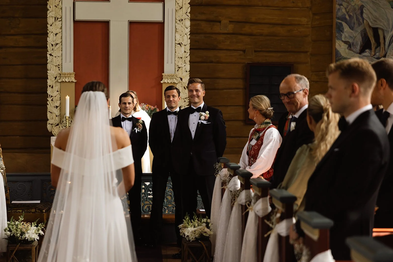 En brud med langt liggende hår i en hvit kjole står foran et alter i en kirke, med fem menn i smokinger og flere kvinner i tradisjonelle klær til høgre.