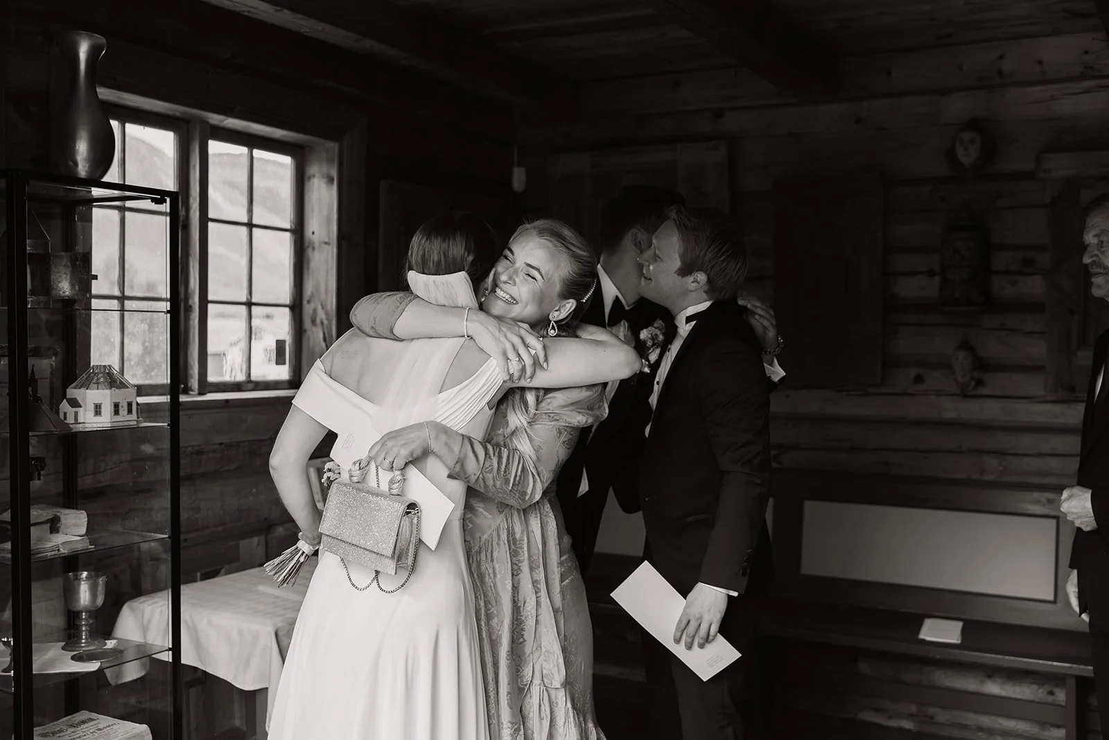 Liten gruppe mennesker som klemmer og vasker at en kvinne i en brudekjole smiler og blir omfavnet på en innendørs feiring.