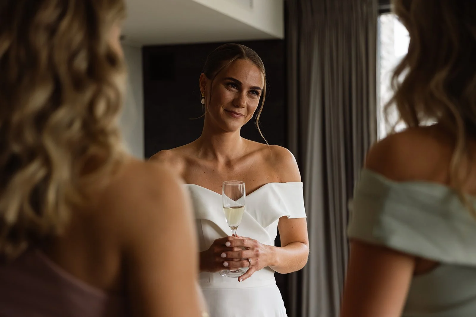 En kvinne i en elegant hvit kjole holder et glass champagne og smiler, omgitt av andre kvinner i festklær.