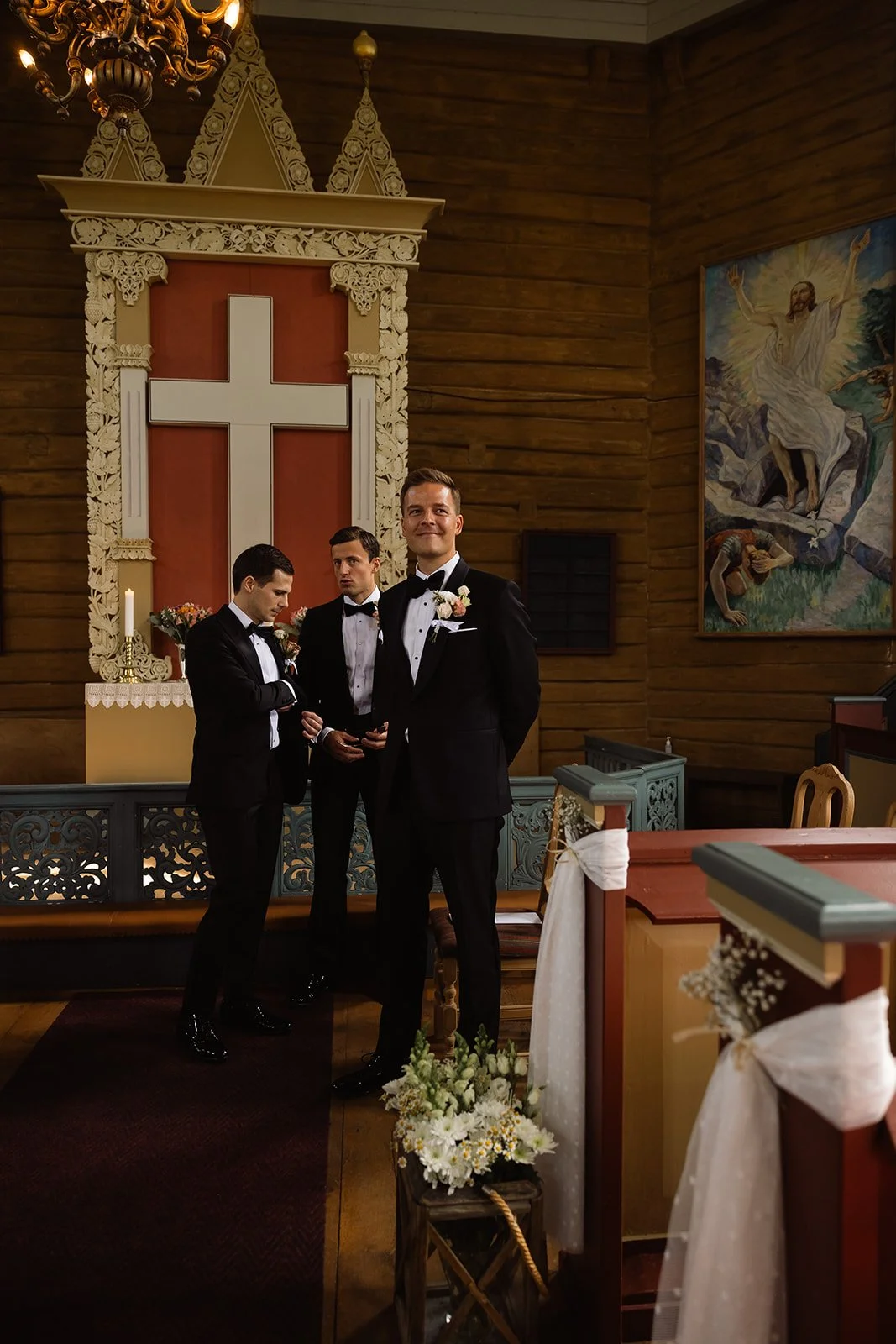 Tre menn i sorte dresser, foran et alter med et kors i en kirke. De er kledd for en bryllupsseremoni, med blomster og hvite buketter. En gutteklær og to voksne menn, kanskje brudgommen og brudeparets forlover. Kirken har trevegger av tre, og det heng