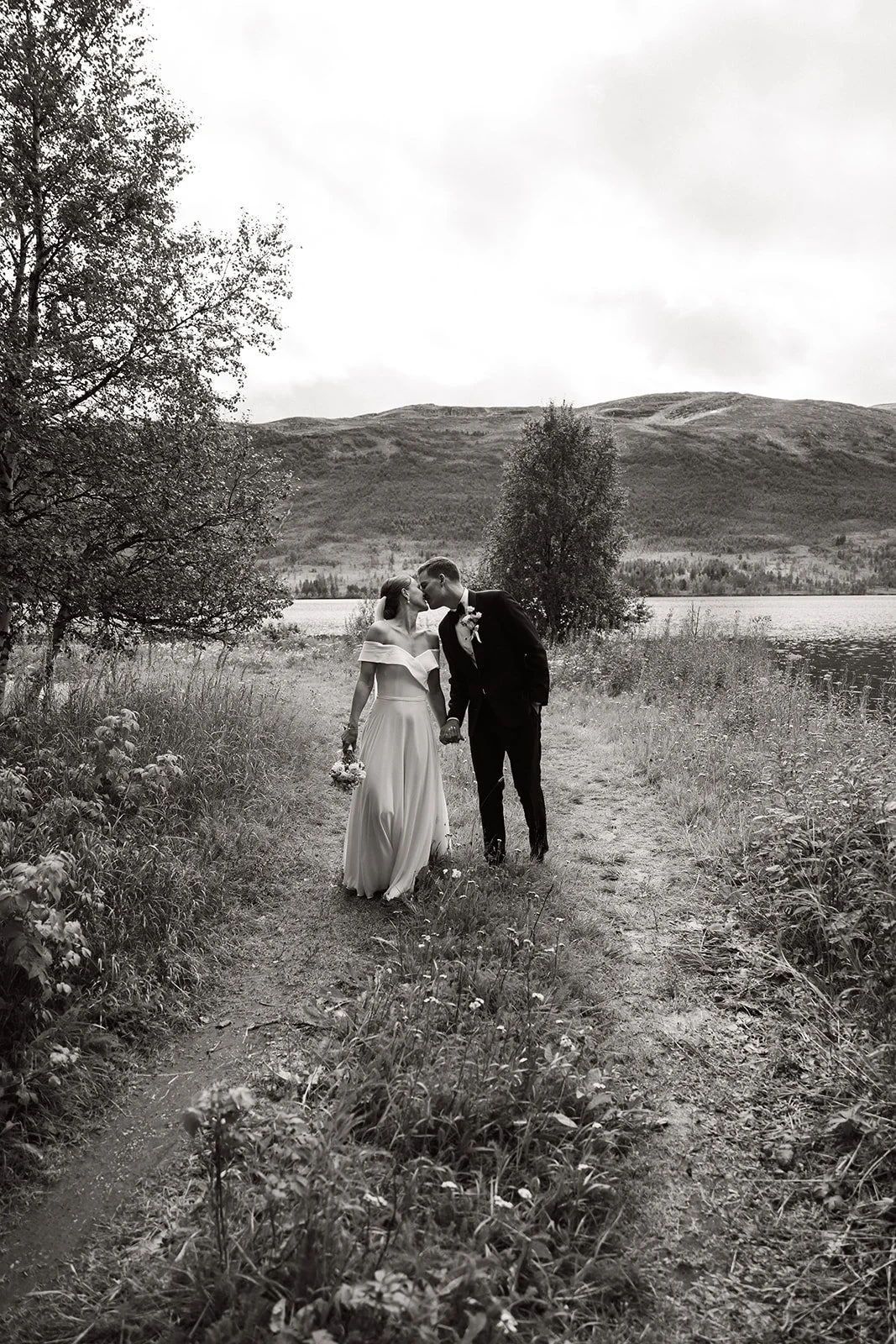 Sort-hvitt bilde av et nygift par som går hånd i hånd langs en sti ved en innsjø med fjell i bakgrunnen, i et naturlig landskap.