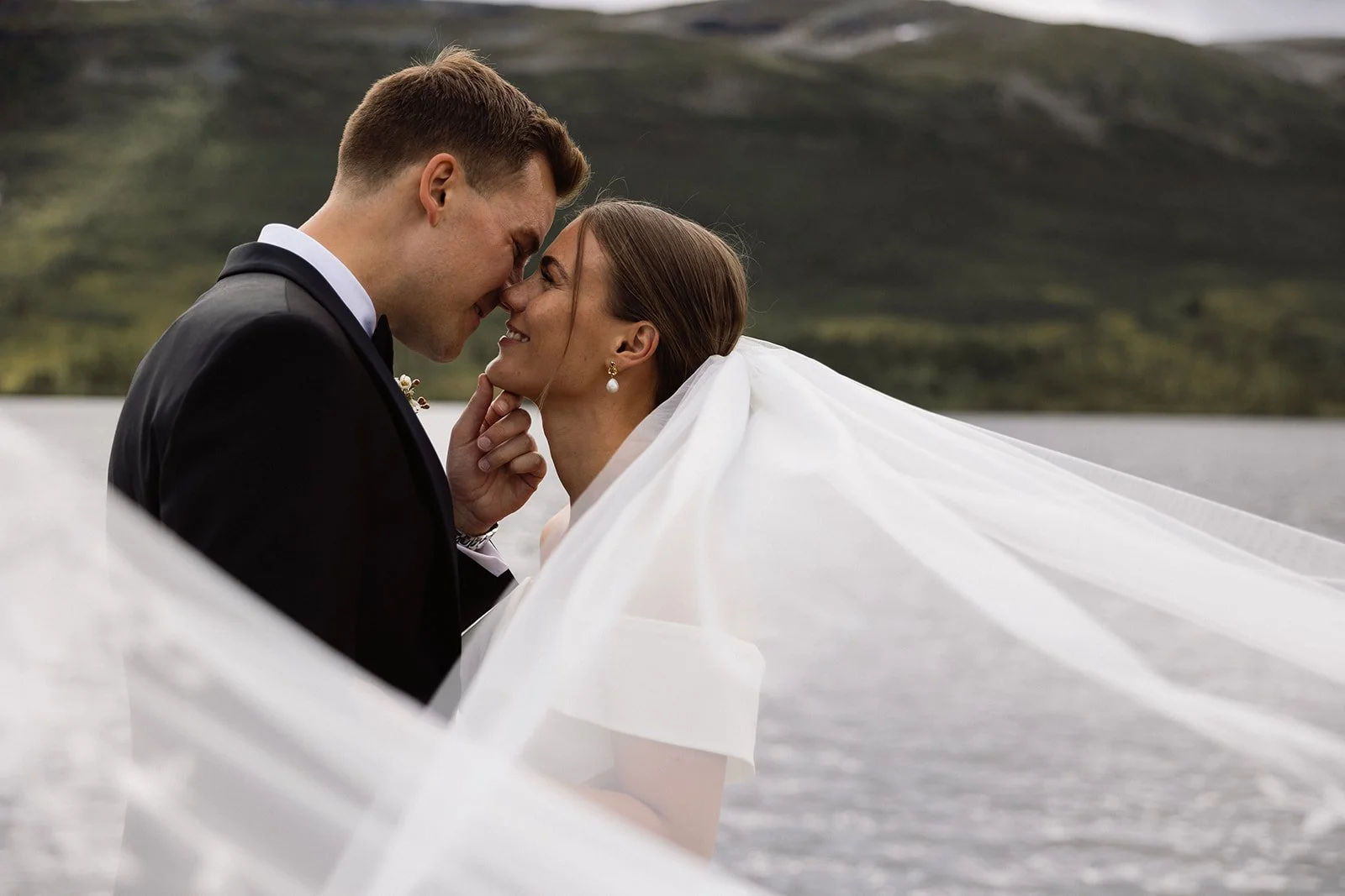 Et brudepar står nært på vannet med fjell i bakgrunnen, og de ler og ser kjærlige ut